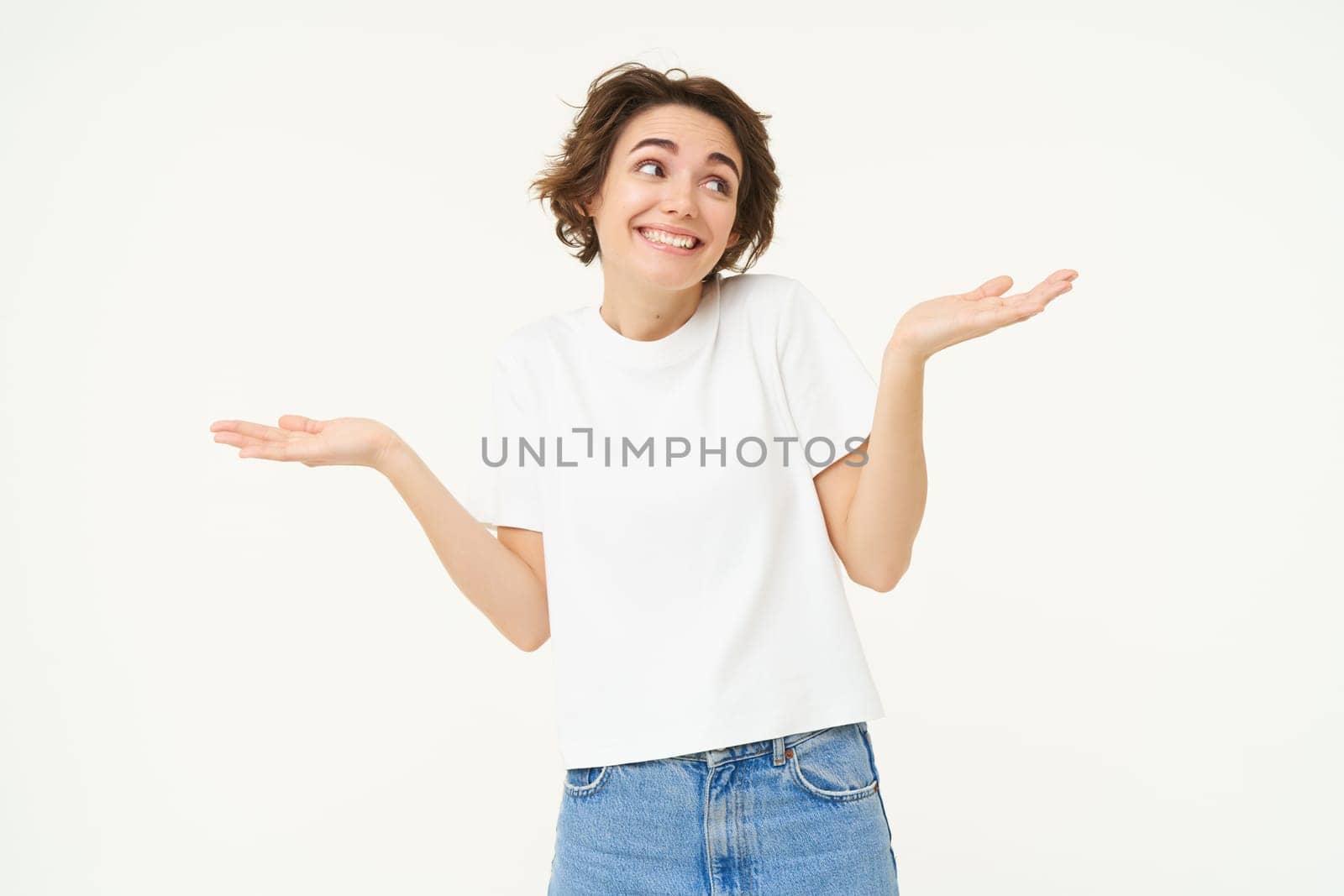 Portrait of girl with short hair, shrugs shoulders, looks unaware, clueless, doesnt know what to do, stands over white studio background.