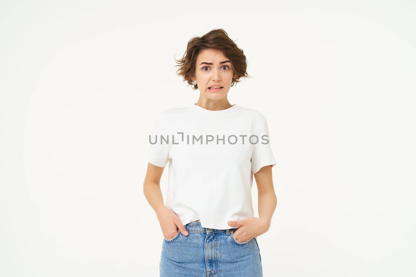 Portrait of girl with concerned face, looks unsure, grimacing, looking confused, standing over white background.