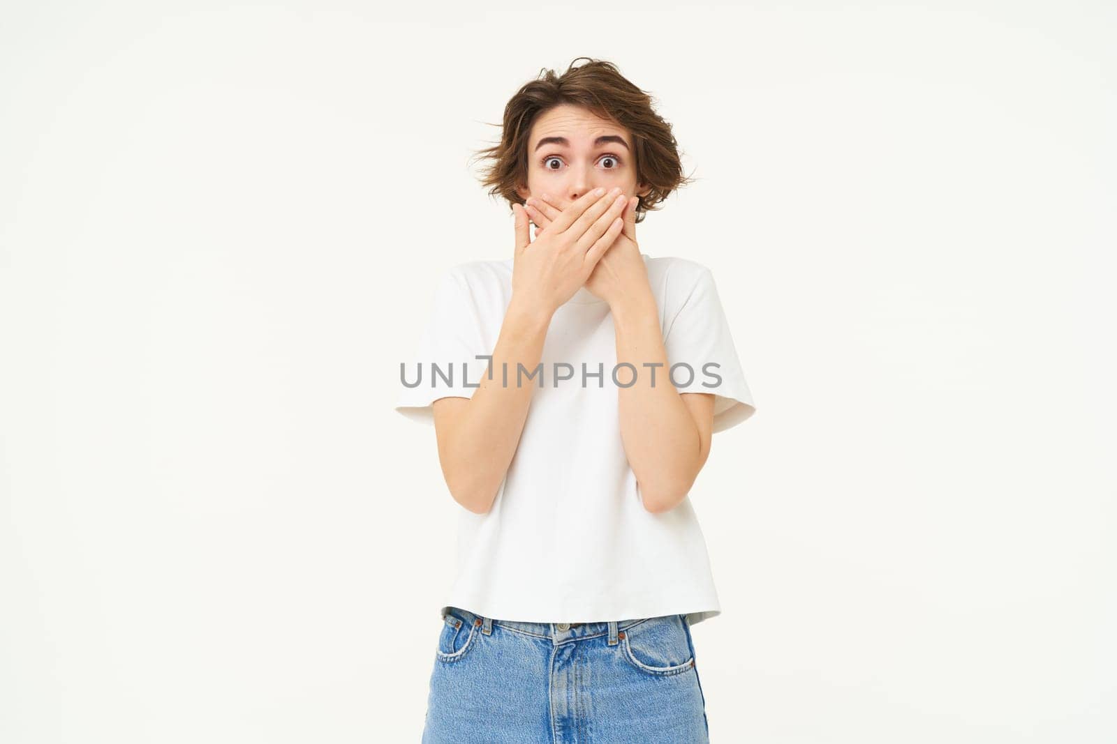 Portrait of woman with surprised face, covers her mouth and gasps shocked, stands isolated over white background.