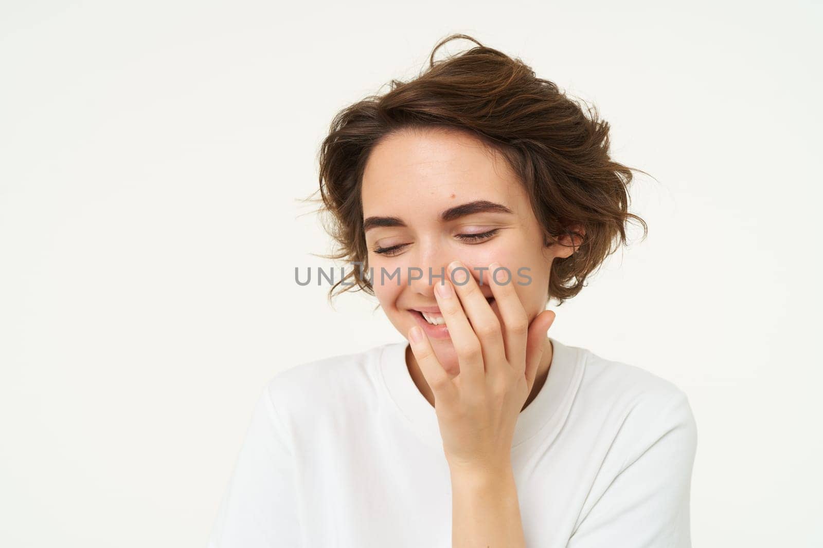 Portrait of brunette woman laughing, smiling and covering face with hand, looking shy and cute, standing over white background. Copy space