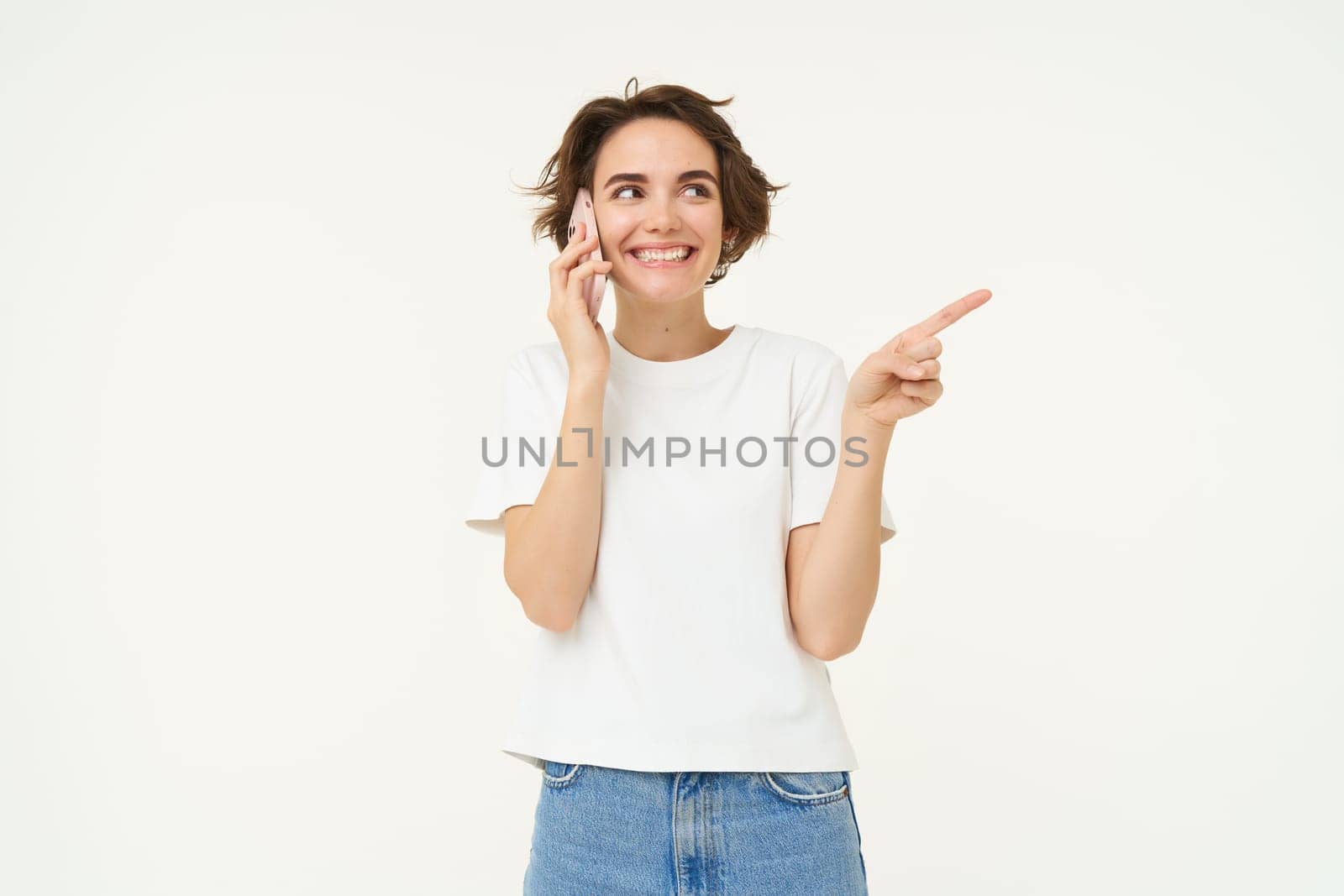 Portrait of friendly, smiling cute girl, pointing finger right, talking on mobile phone, showing smth aside on white copy space, isolated on studio background.