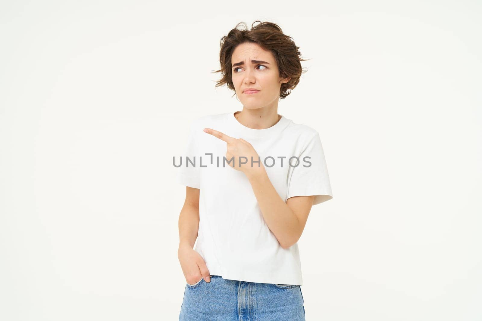 Portrait of woman looking with doubt, pointing left, staring at something with unsure, concerned face expression, standing over white studio background.