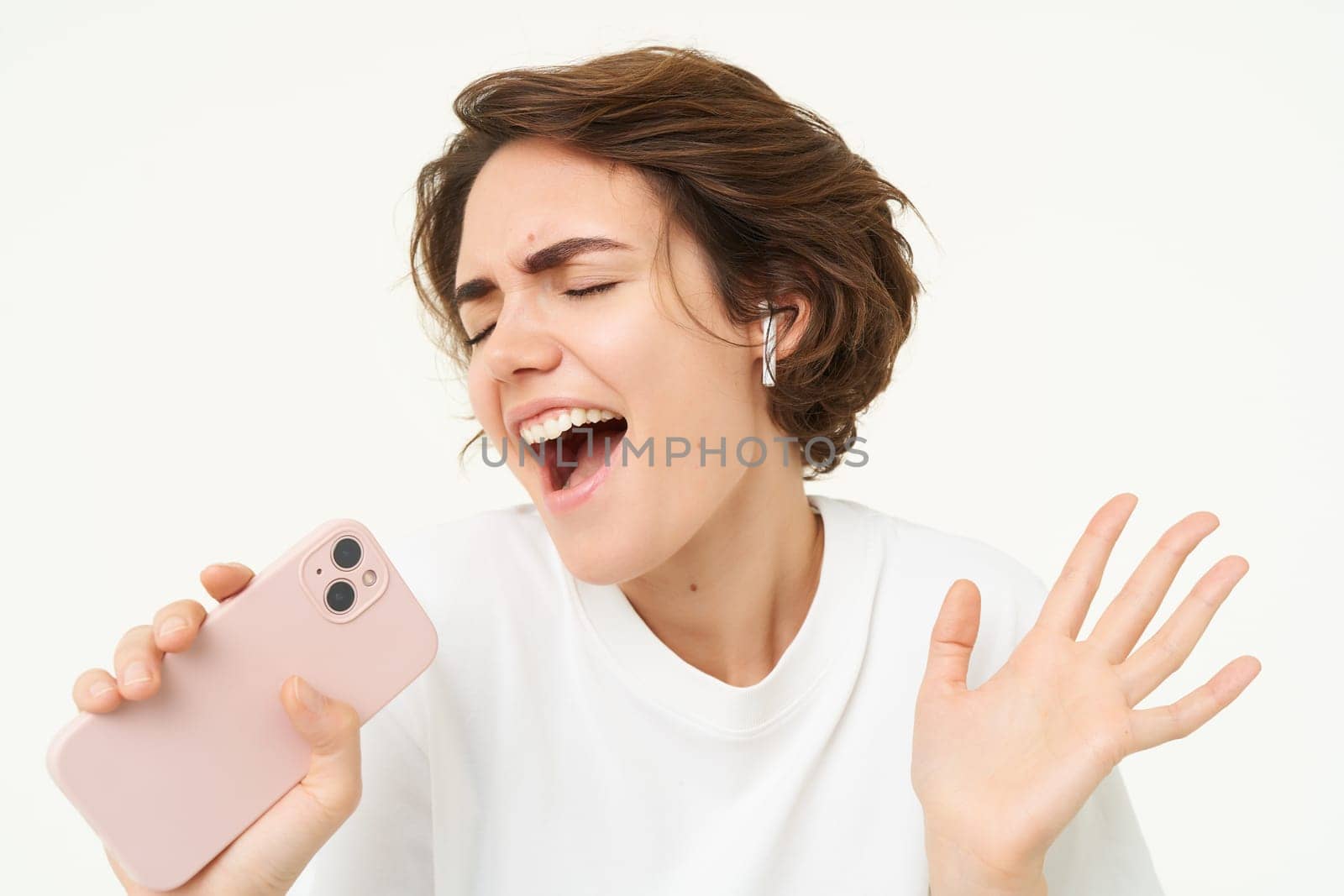 Woman singing along, listening music in wireless headphones, holding smartphone in hand, standing over white background.