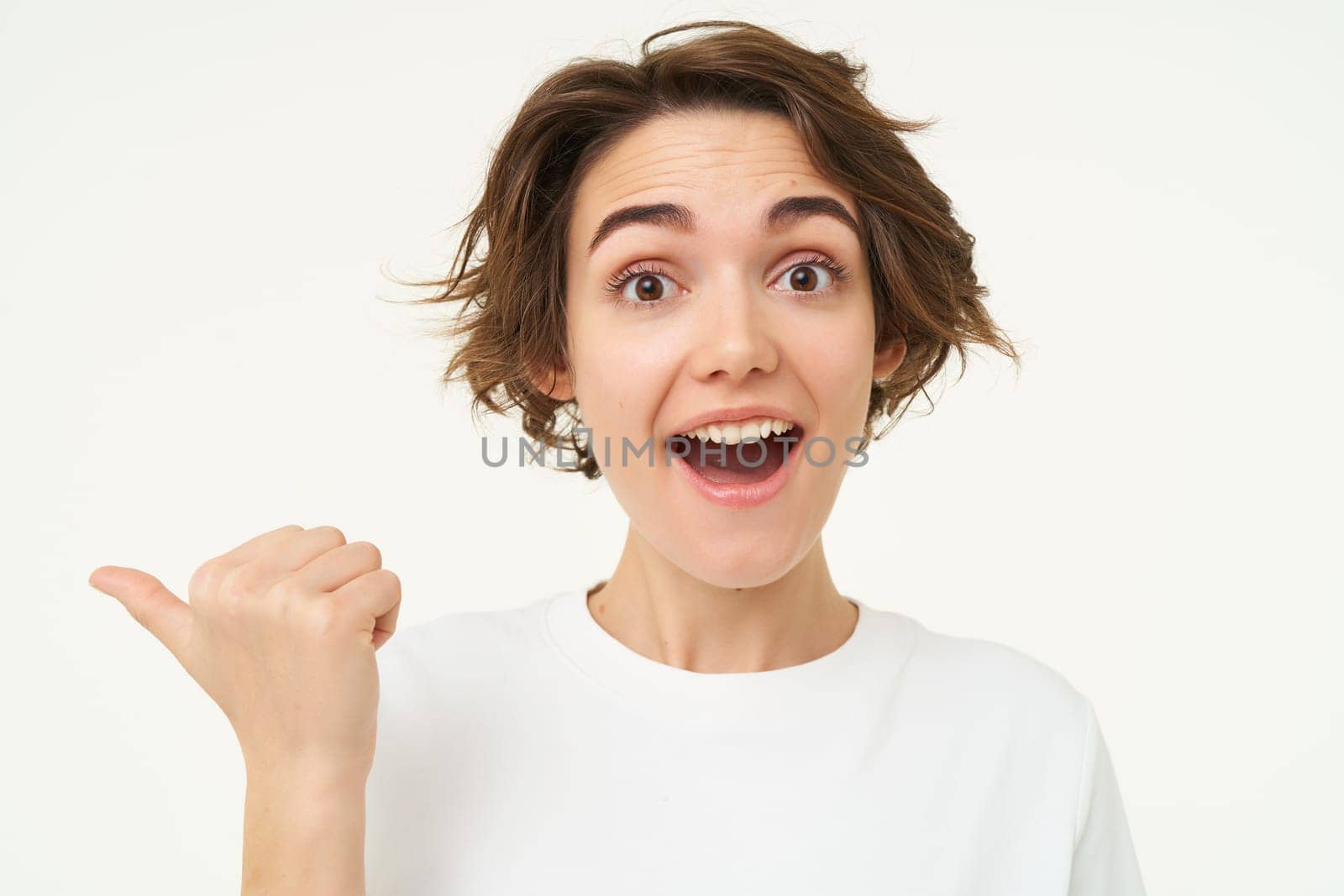 Close up portrait of brunette girl gasps and looks surprised, points left to inquire about promotion, stands over white background.