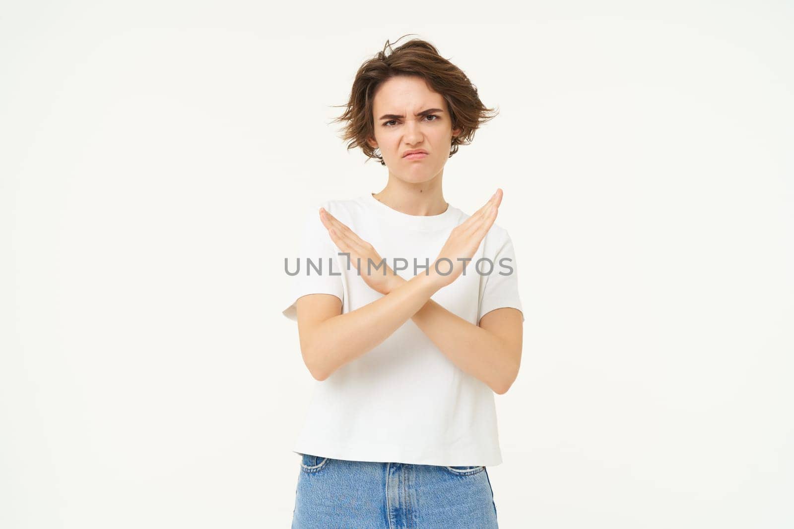 Portrait of woman with serious face, shows cross, stop gesture, prohibit something bad, standing over white background. copy space