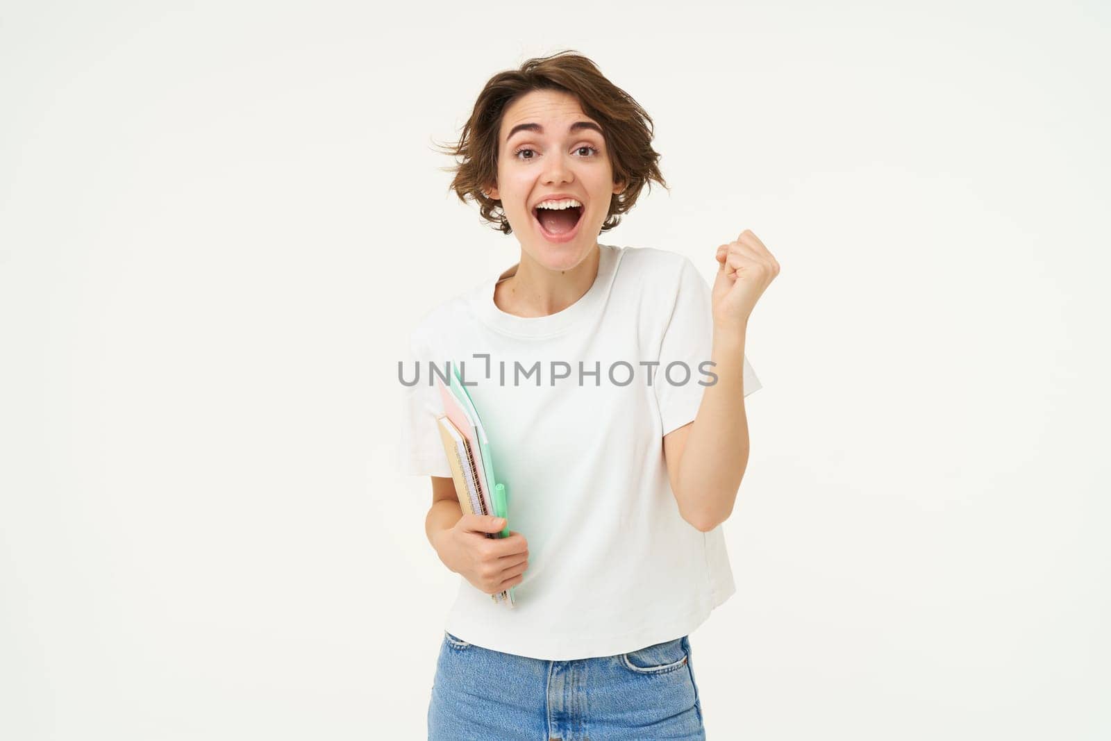 Enthusiastic brunette woman makes fist pump, holds documents and notes, looks thrilled and happy, winning, triumphing, posing over white background.
