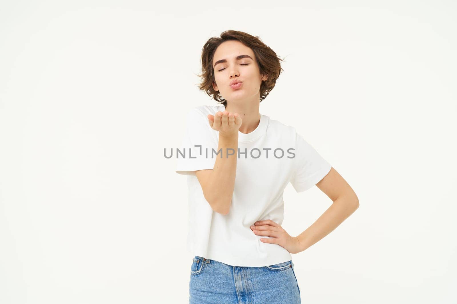 Portrait of young woman sending air kiss, blowing at camera and smiling, standing over white background.