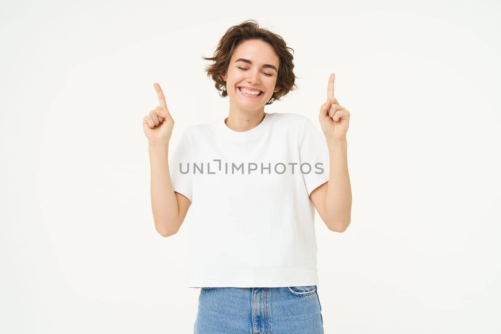 Image of happy, beautiful modern woman, pointing fingers up, showing advertisement on top, smiling, standing over white background.