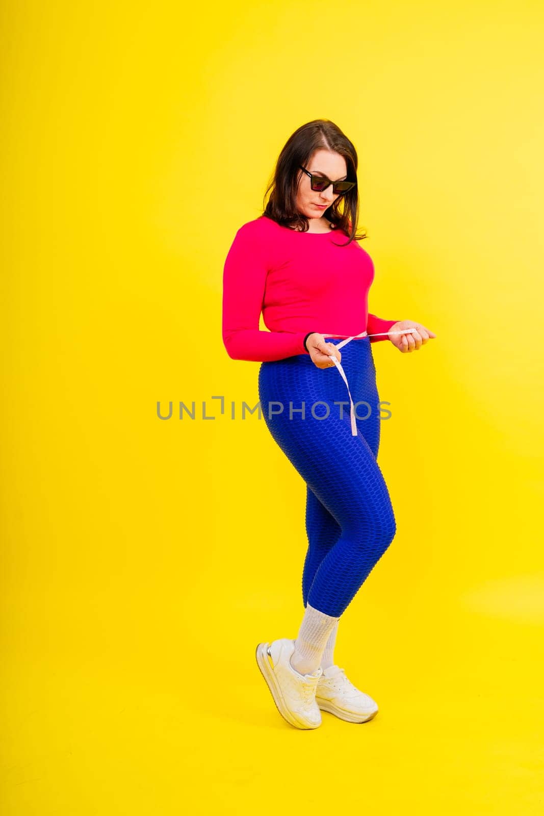 Overweight woman measuring waist before weight loss in studio shot on yellow background by Zelenin