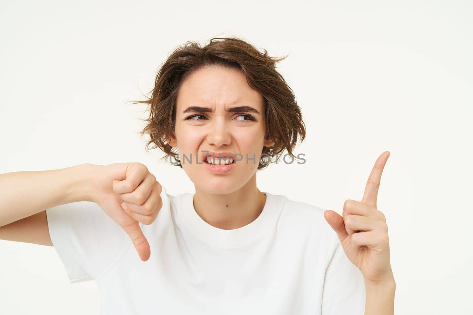 Portrait of disappointed girl, shows thumbs down, pointing at bad product, dislike something, standing over white background.