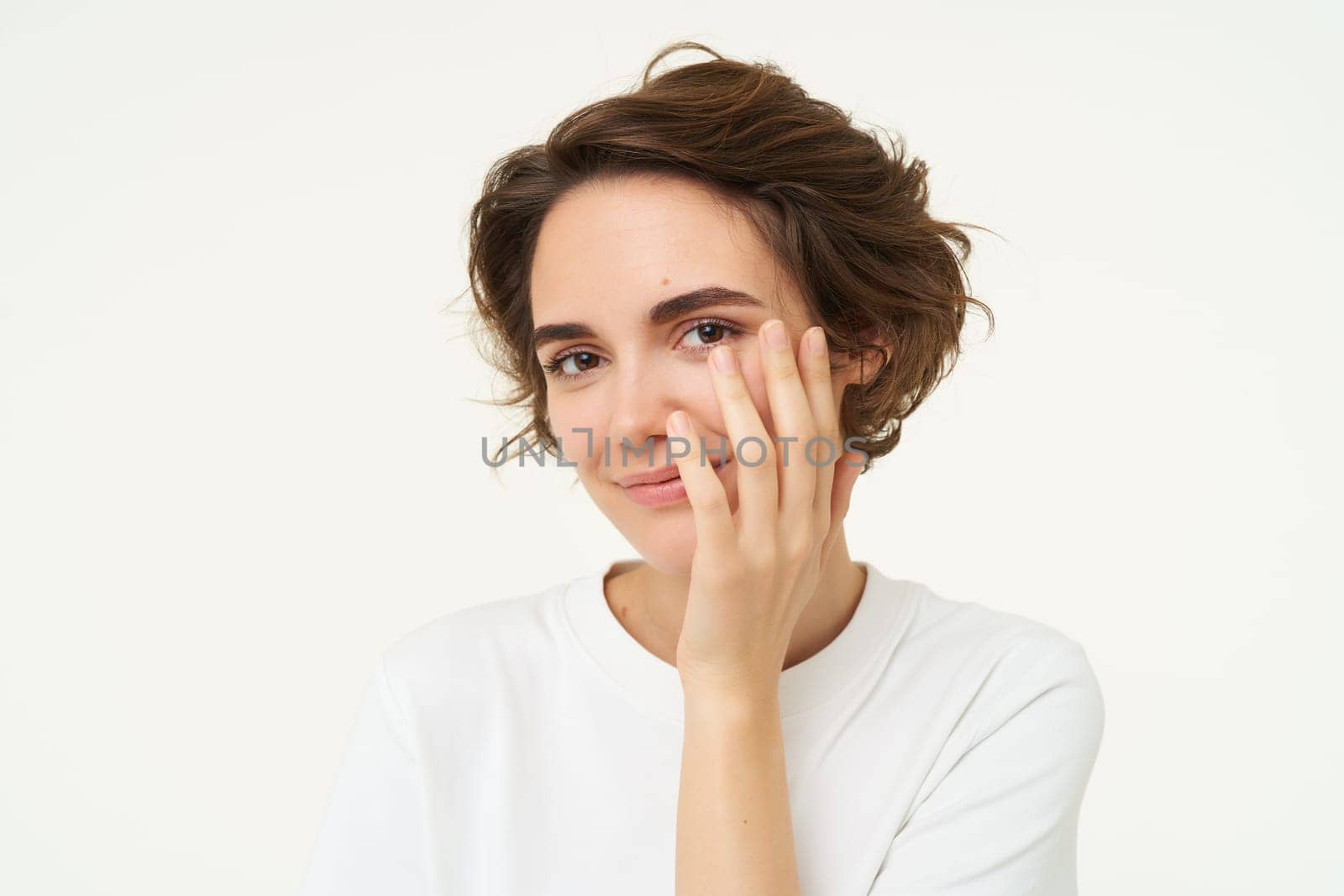 Close up of brunette woman looks shy, covers face with hand and smiles, gazing flirty at camera, stands over white background.