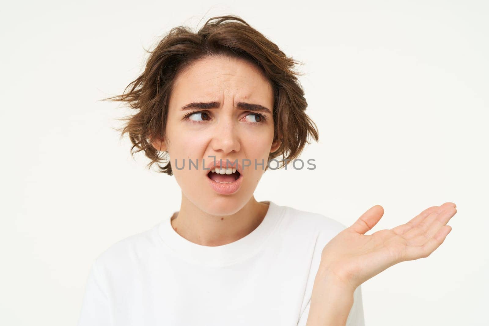 Portrait of disappointed young woman complaining, holding hand sideways, talking, expresses dislike and irritation, standing over white background.