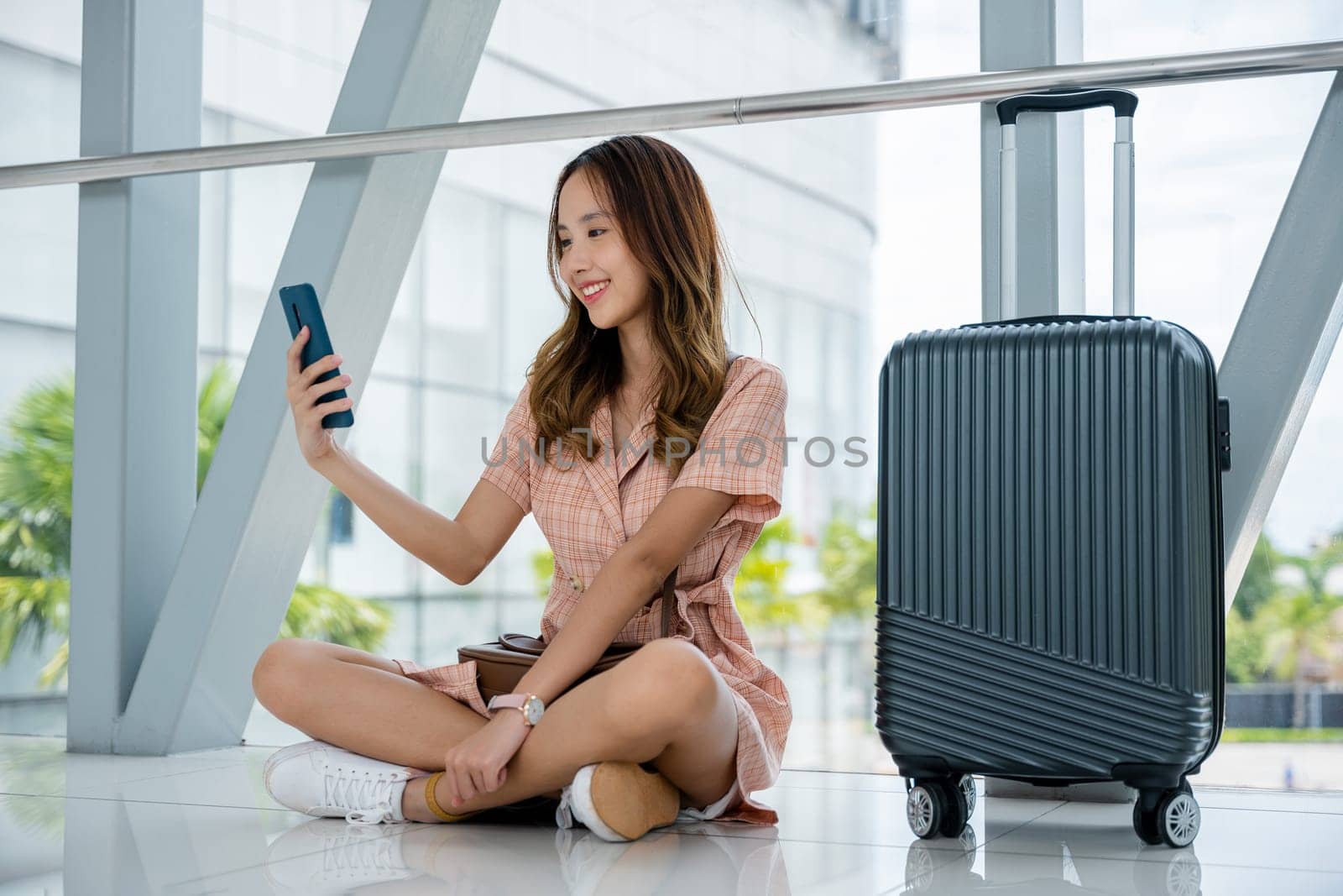 Traveler using her smartphone to check flight while sitting with her luggage on the airport floor by Sorapop