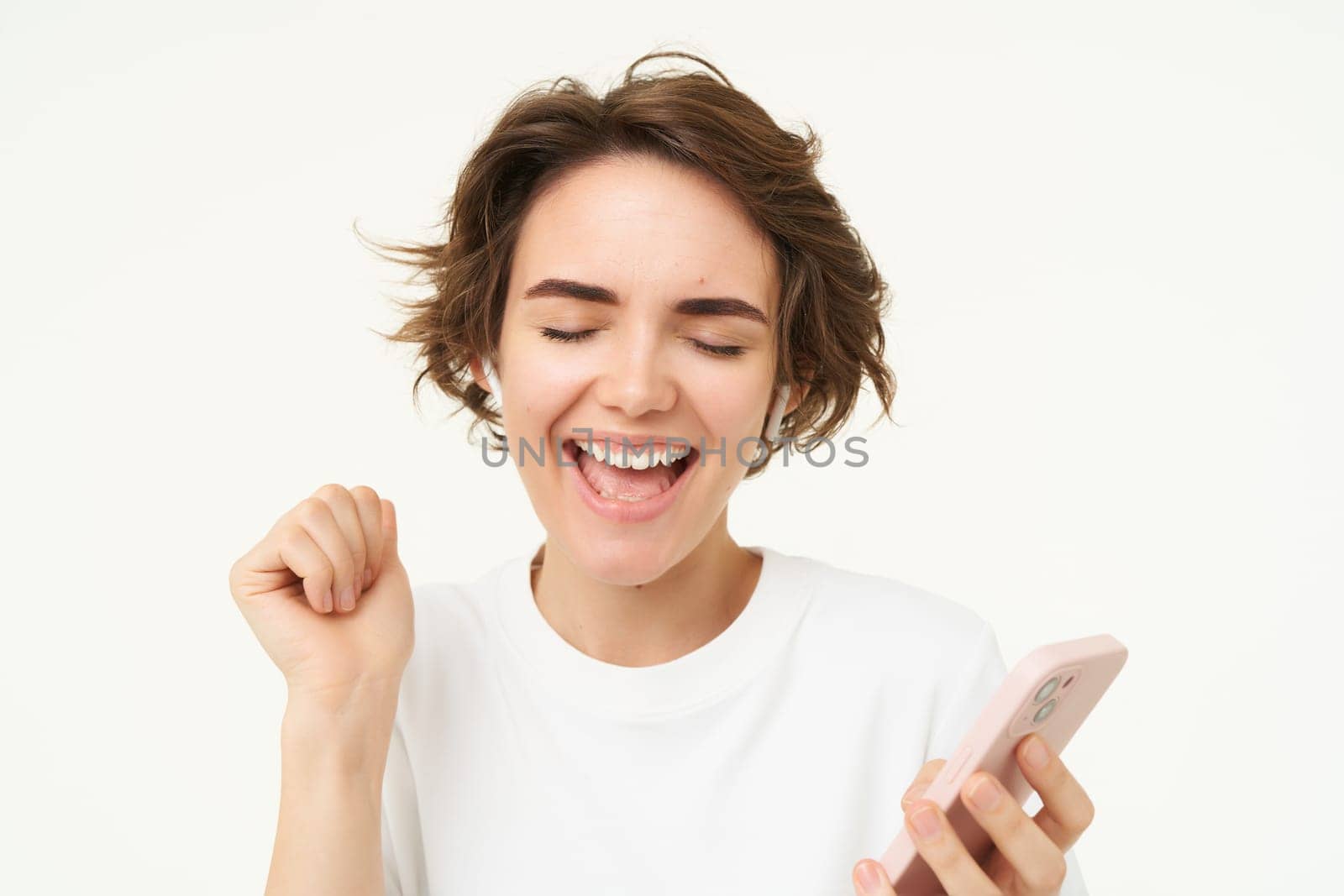 Woman singing along, listening music in wireless headphones, holding smartphone in hand, standing over white background.