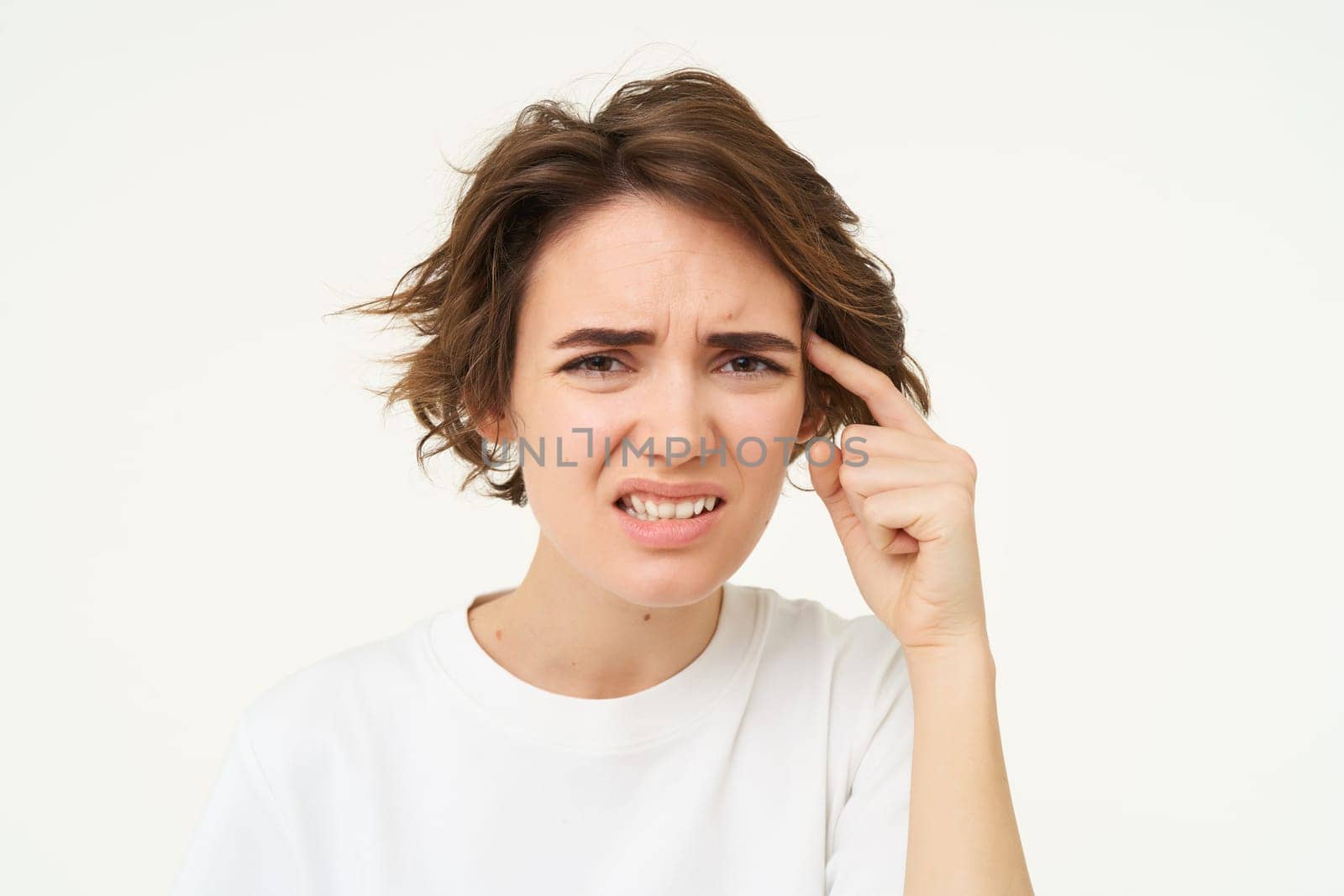 Portrait of annoyed brunette woman pointing finger at head, frowning and grimacing, thinks someone is stupid, standing over white background. Copy space