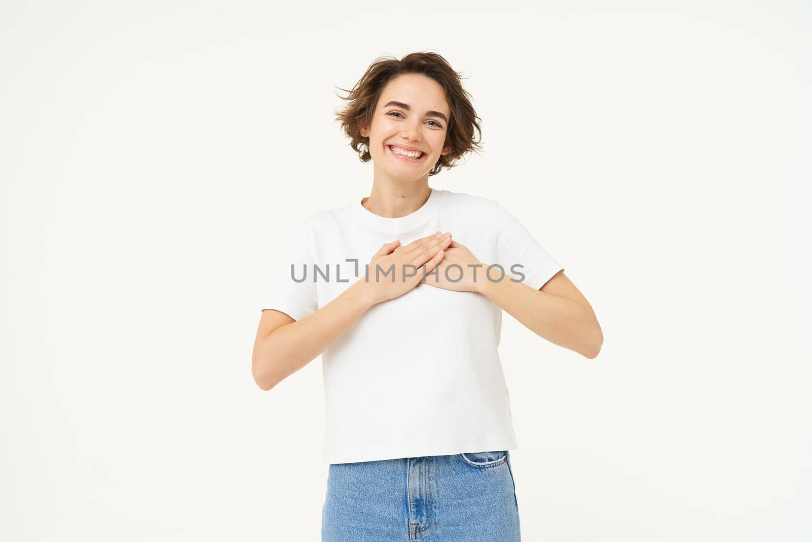 Portrait of woman expressing care, self-love and caring emotions, holds hands on chest, looking with smile at camera, isolated over white background.