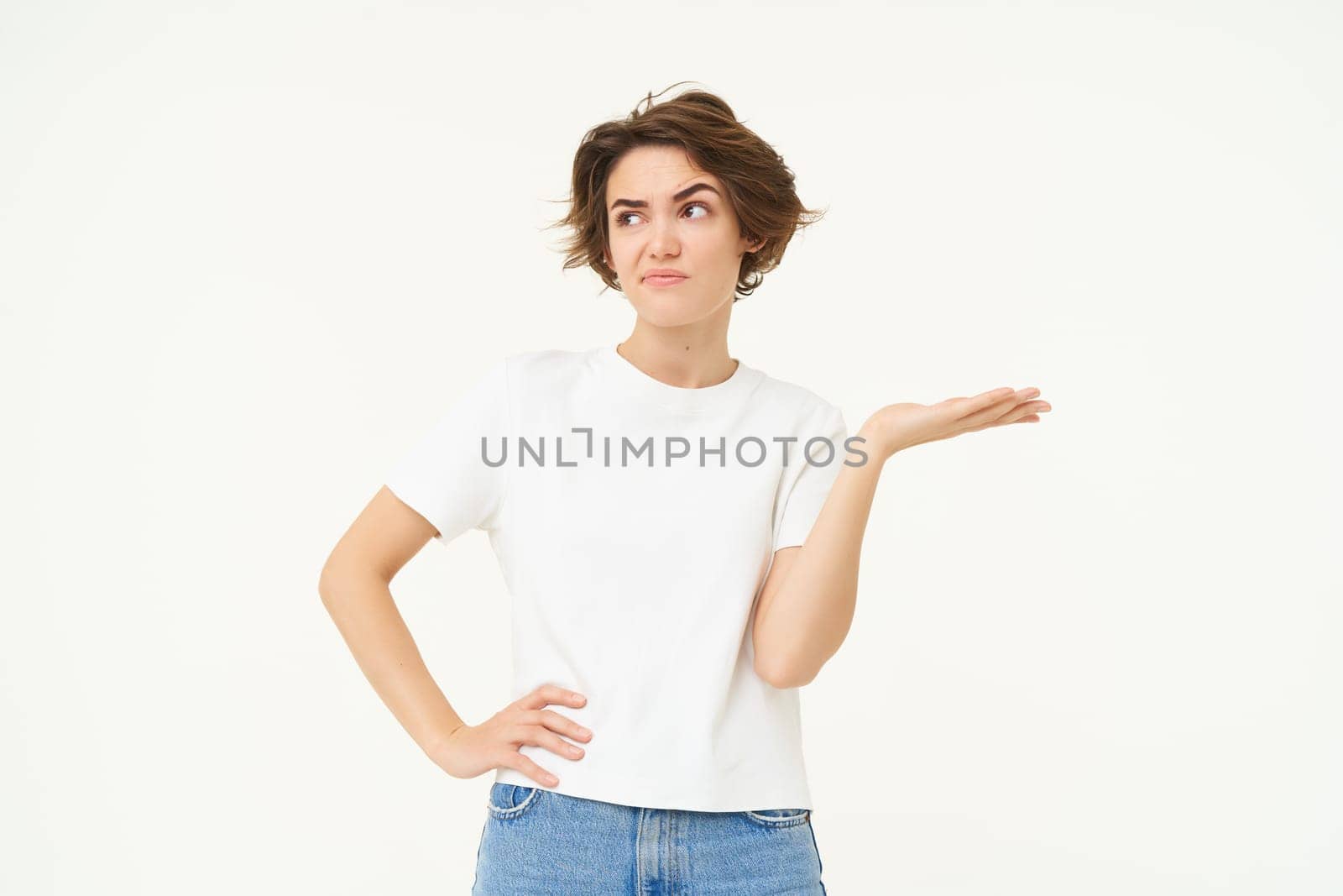 So what. Frustrated, upset young woman, raises one hand and shrugs, looking confused, cant understand something, standing over white background.