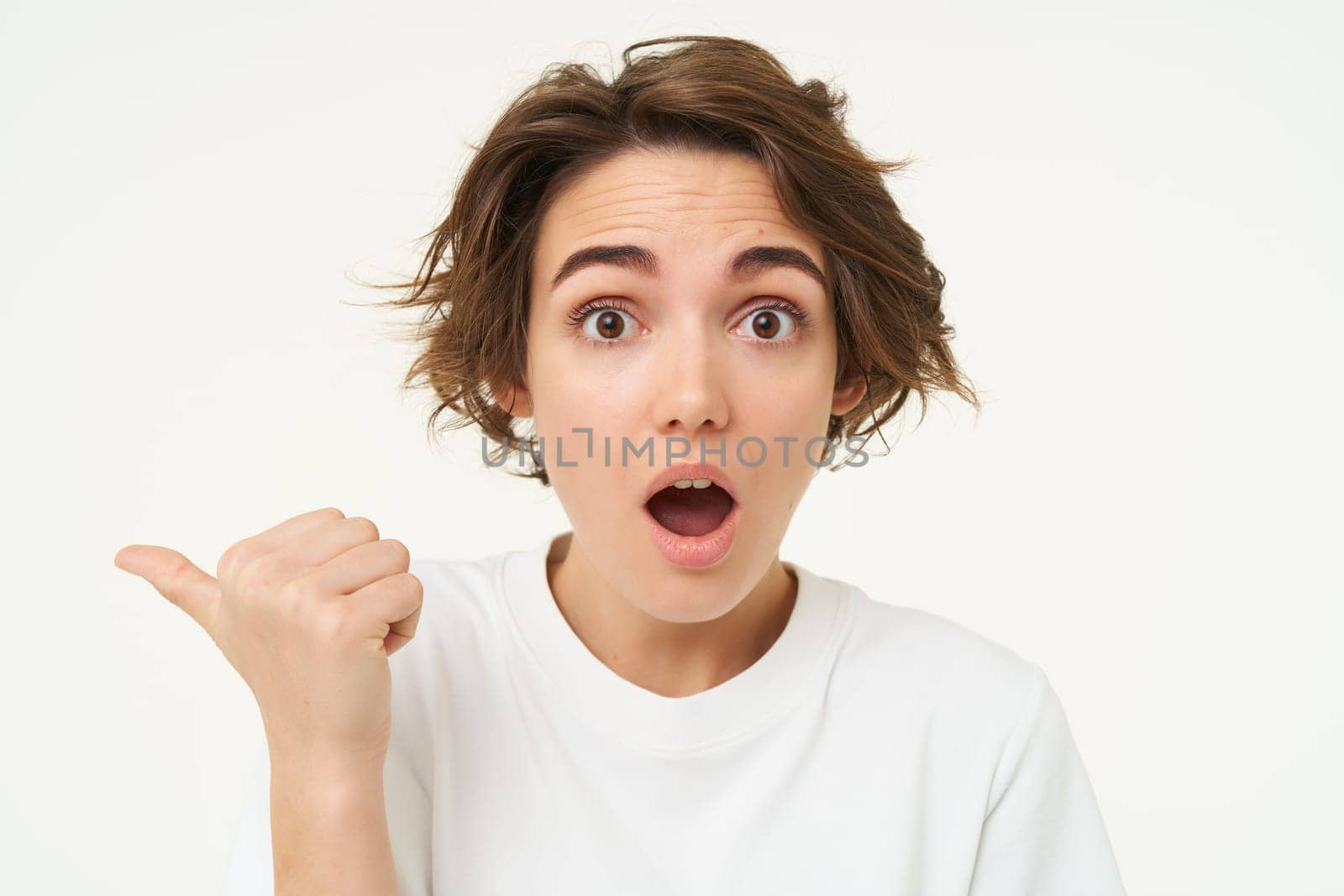 Close up portrait of brunette girl gasps and looks surprised, points left to inquire about promotion, stands over white background.