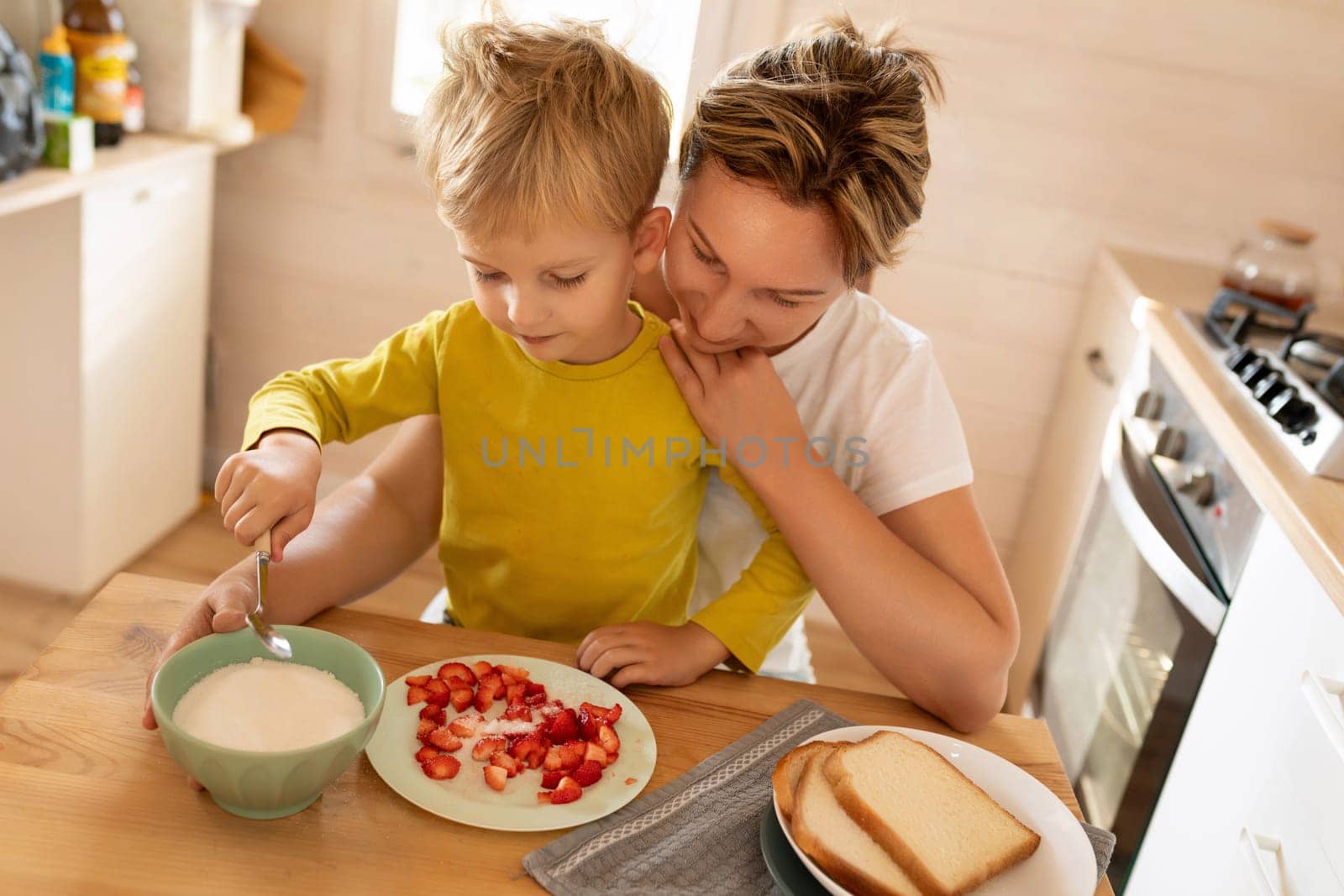 little Caucasian blond son and mother preparing breakfast with strawberries while sitting in the kitchen.