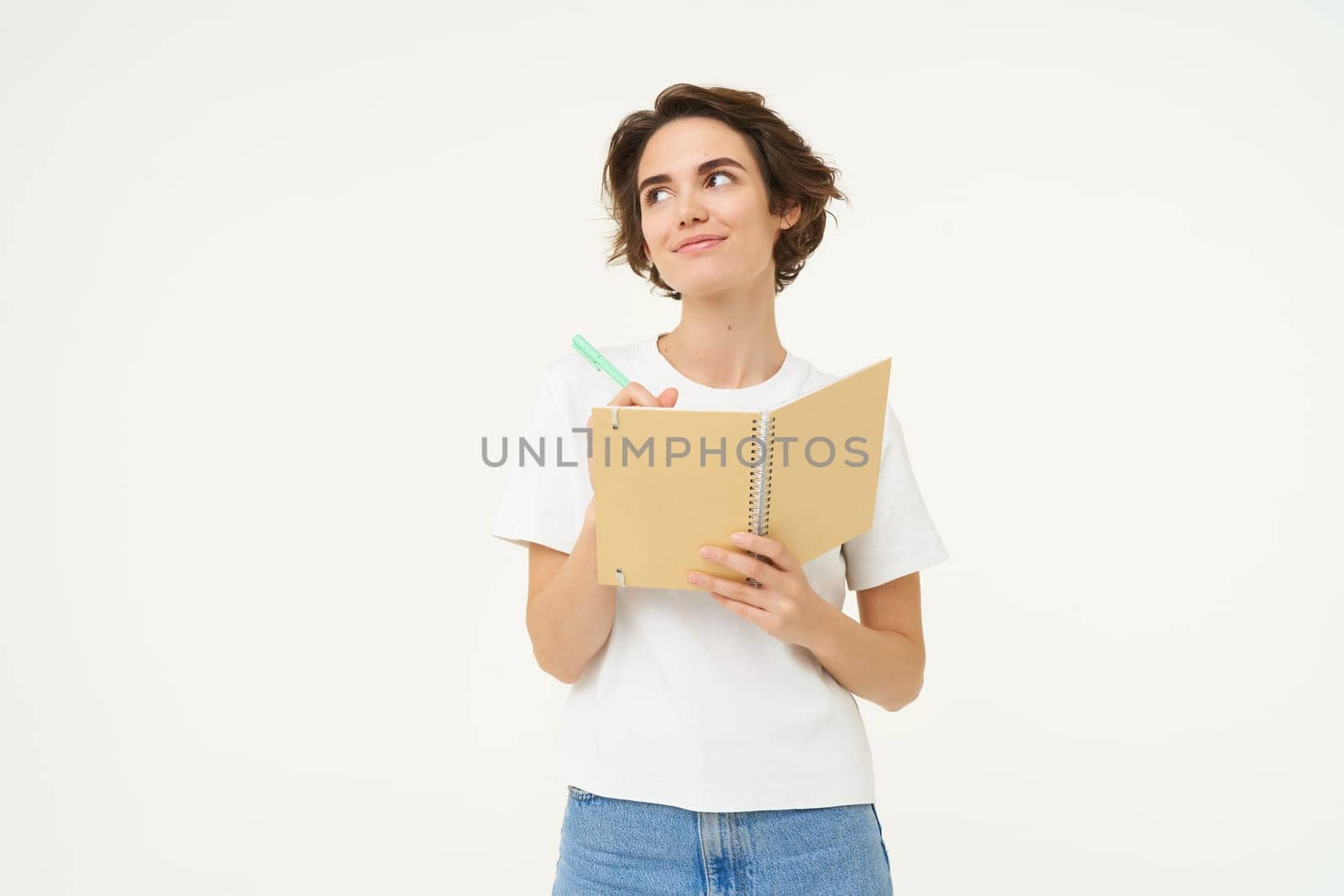 Portrait of thoughtful and creative woman writing down her thoughts in diary, doing homework, looking aside and thinking, posing over white background.