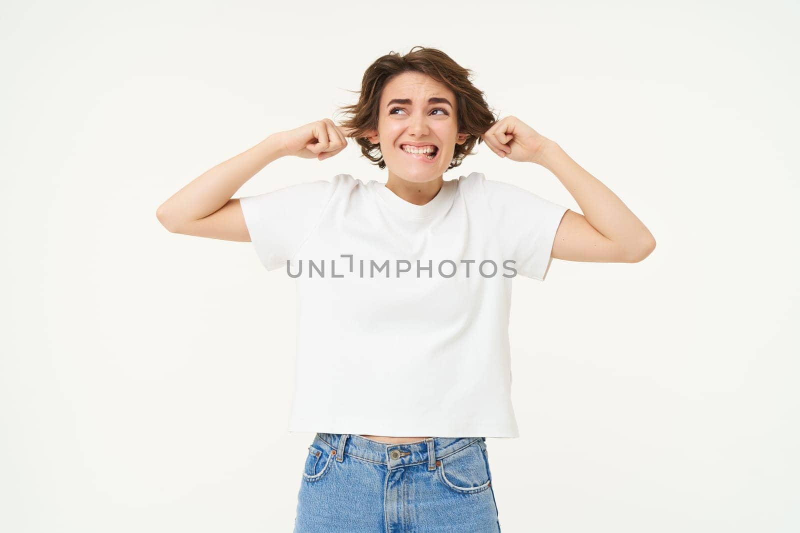 Woman standing annoyed, holding fingers in ears, unwilling to listen, feeling discomfort from loud noise, standing over white background.