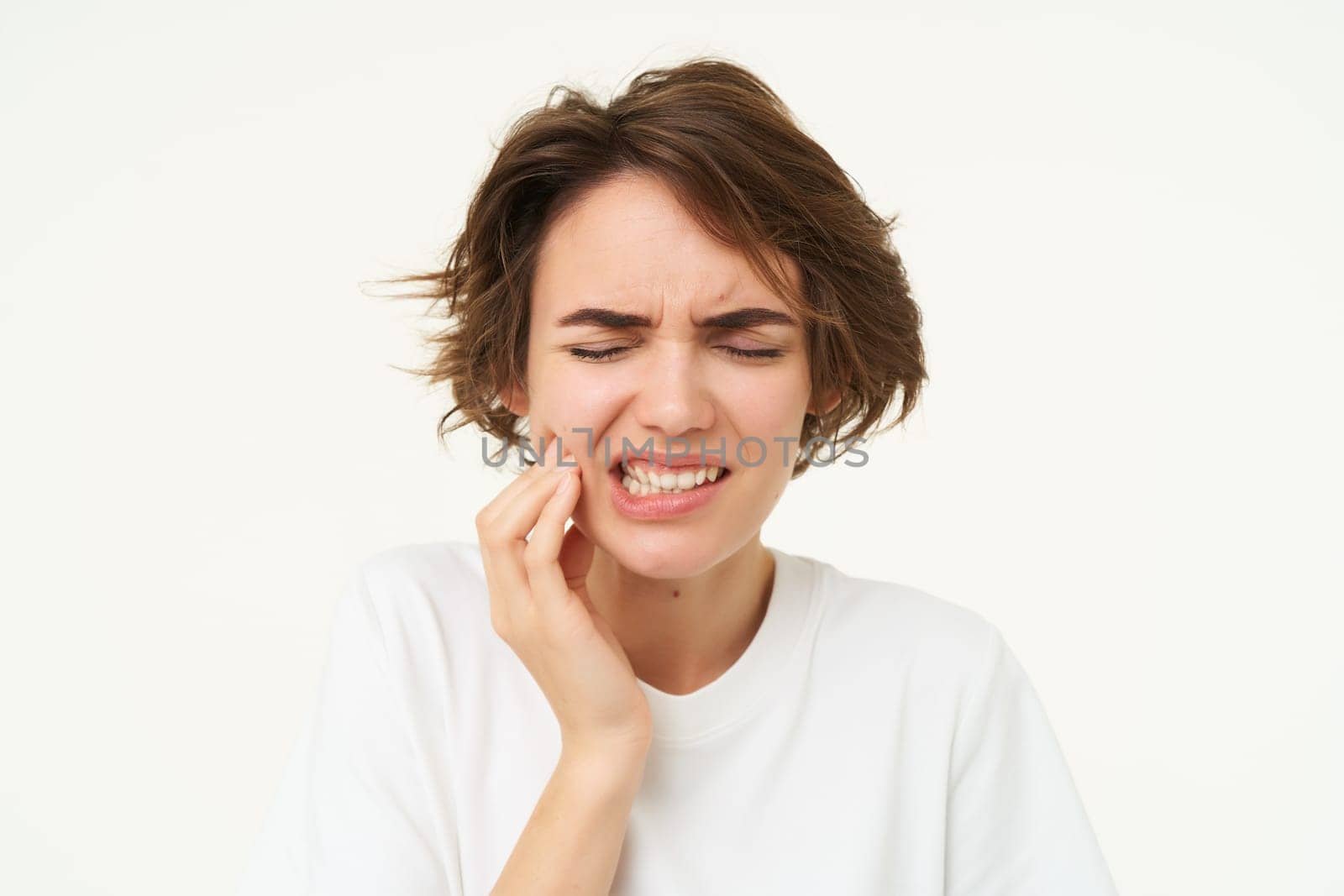 Close up of brunette woman touches her tooth, has a toothache, dental problem, painful discomfort in mouth, stands over white background.