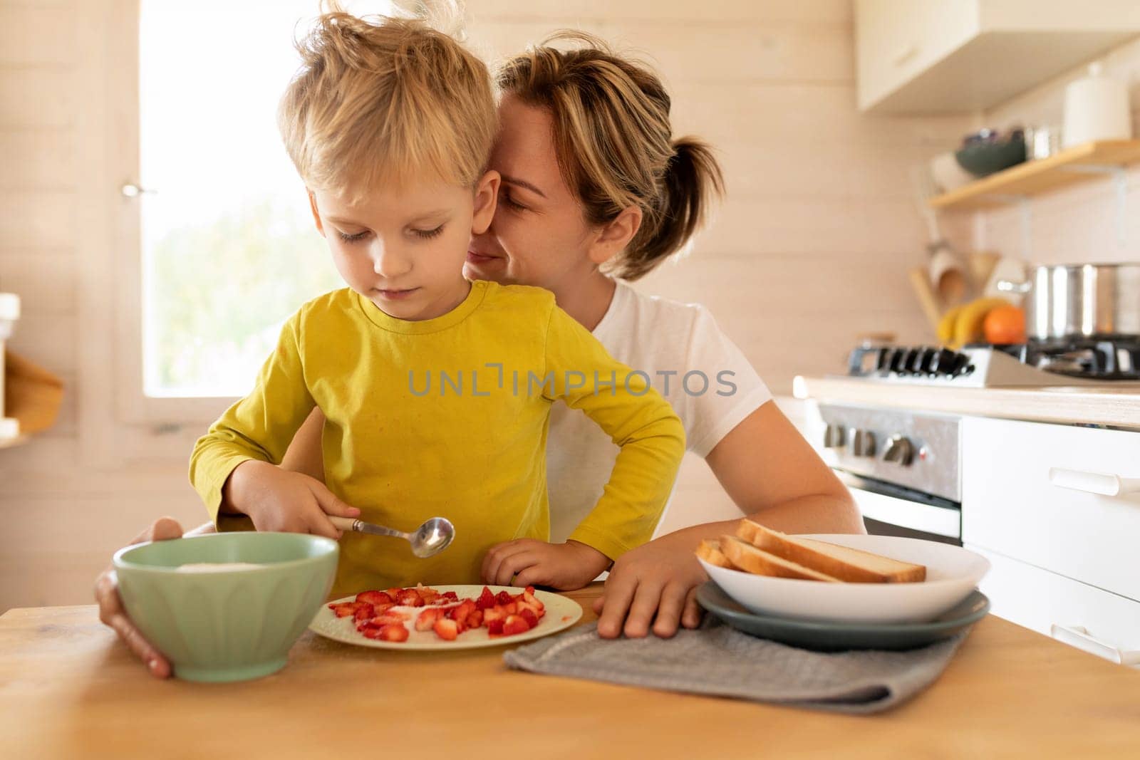 rested Caucasian mother hugs her baby boy while sitting in the kitchen by TRMK