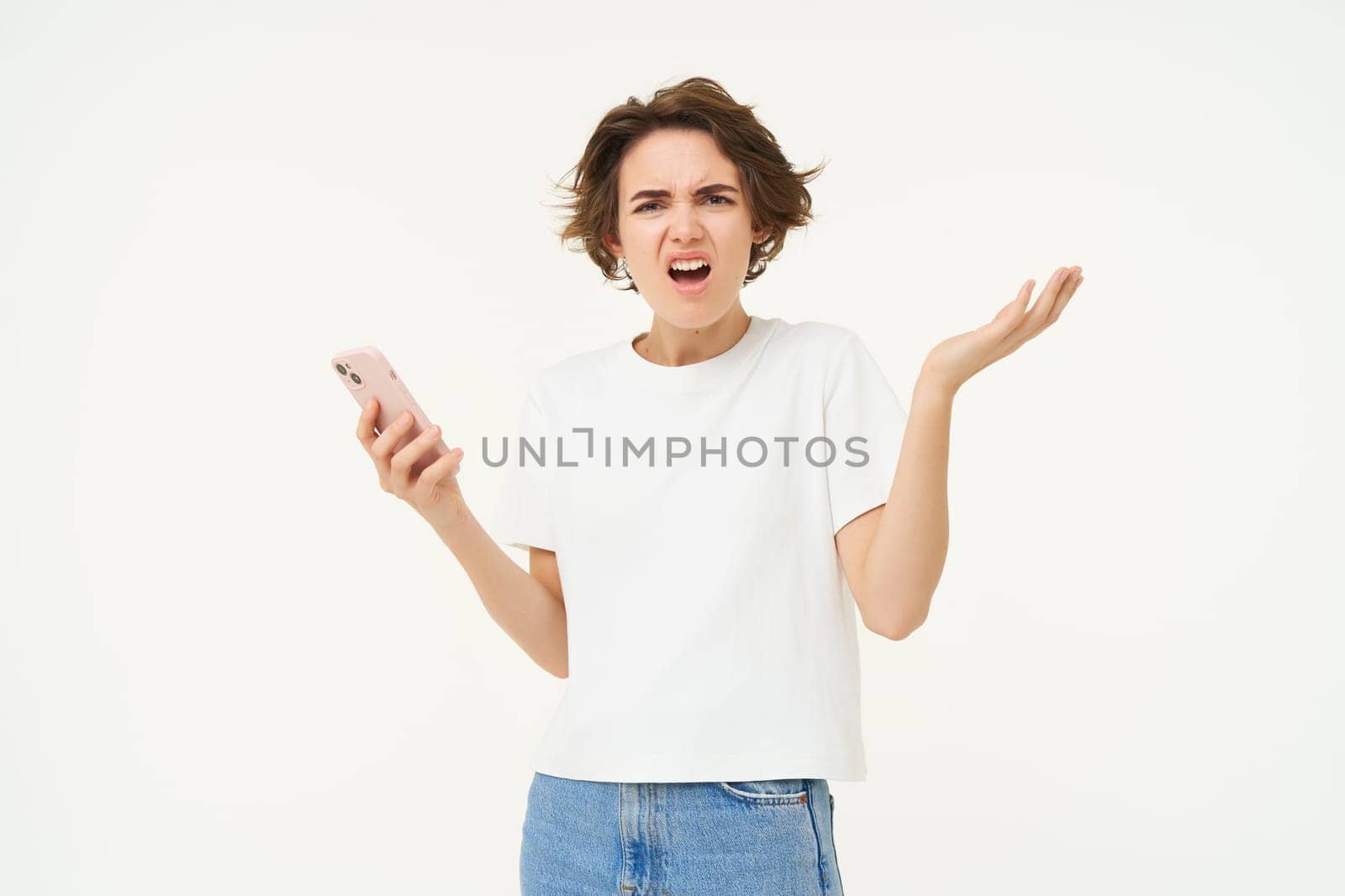 Portrait of confused and upset young woman, holding smartphone, shrugging shoulders and complaining at something online, standing over white background.