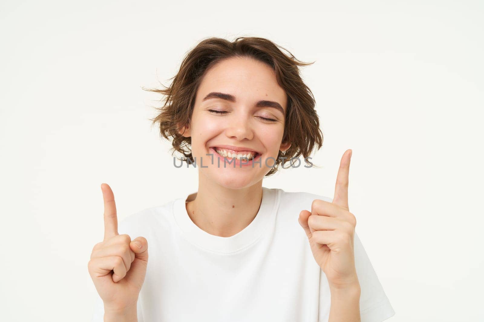 Close up portrait of happy, smiling brunette girl, pointing fingers up, laughing, showing advertisement, standing over white studio background. copy space