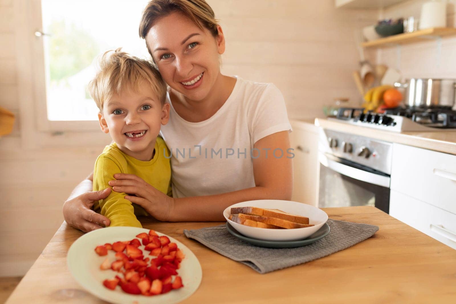 young caucasian blond mother and child son sitting in the morning in the kitchen and preparing breakfast with toast and strawberries.