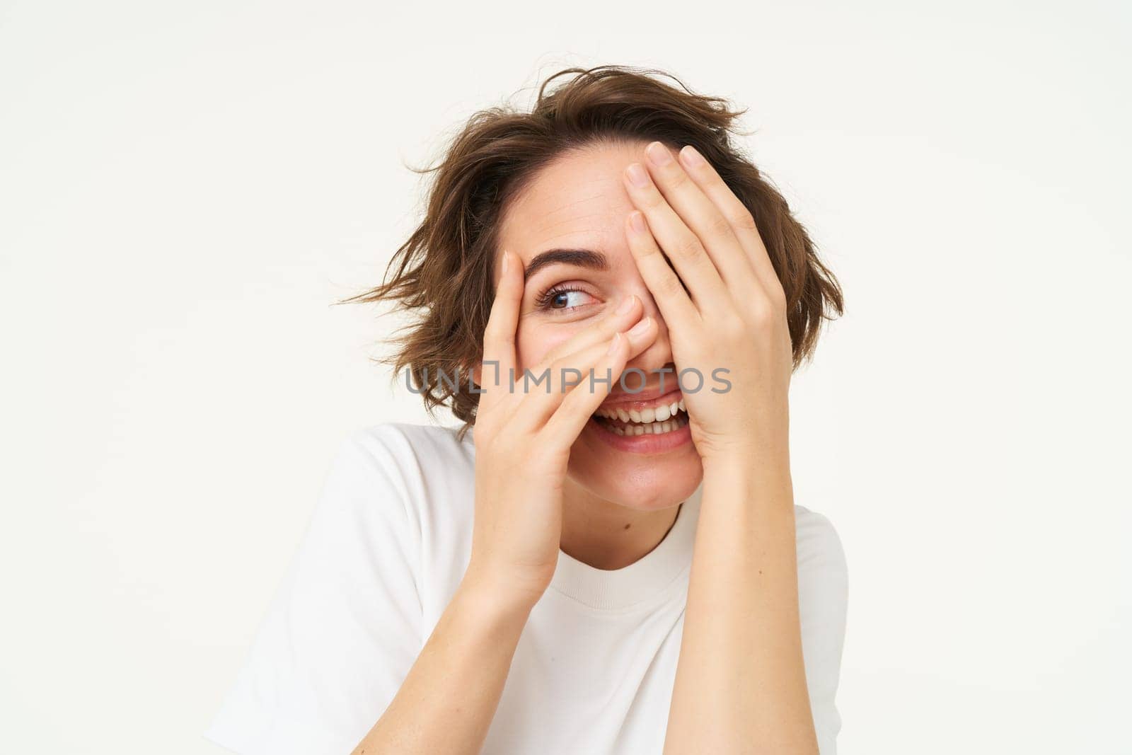 Image of funny woman peeking through fingers, looking at something with curious face expression, waiting for surprise and smiling excited, isolated over white background.