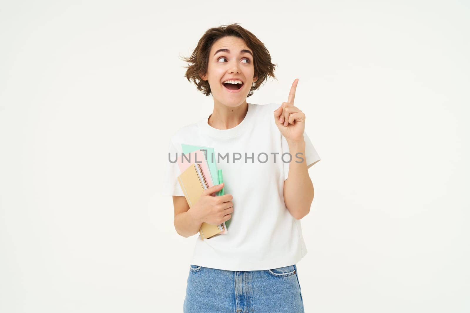 Portrait of brunette woman laughing, student with notebooks pointing at upper right corner, showing banner or advertisement, standing over white studio background.