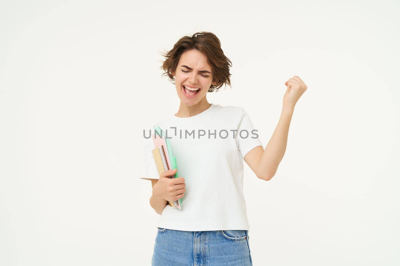 Excited and enthusiastic girl, feeling thrilled, makes fist pump and screams from joy, posing with notebook against white studio background.