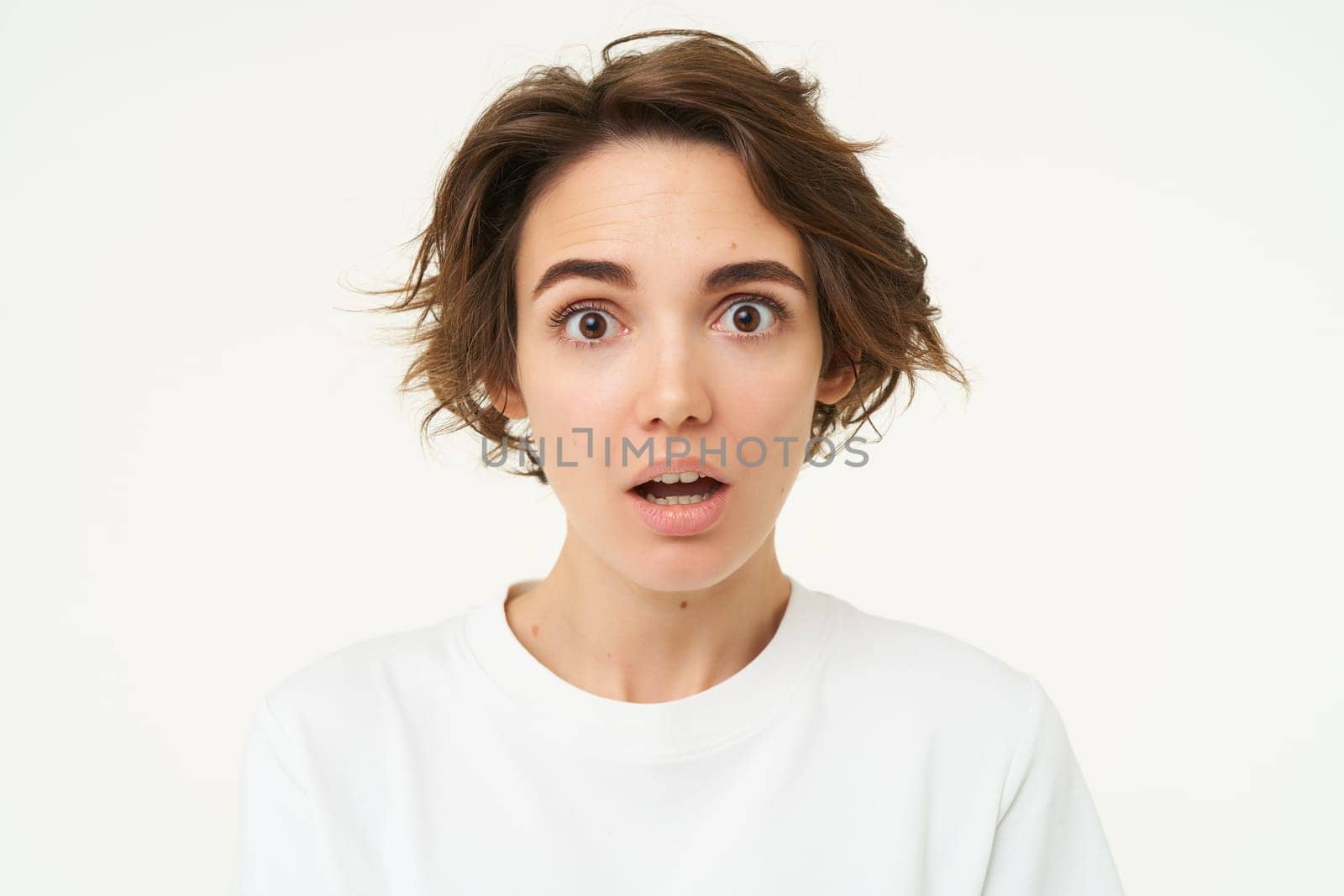 Close up shot of woman with shocked face, gasping and staring startled at camera, confused and surprised by something, standing over white background.