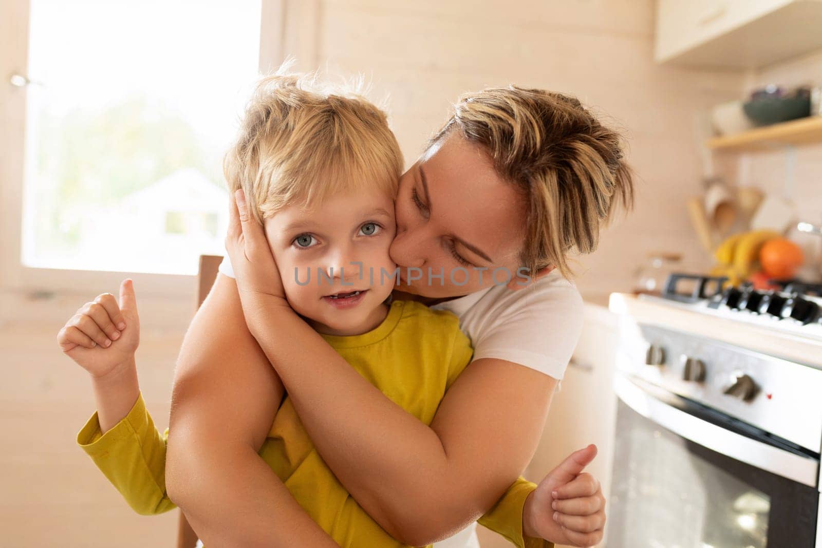 rested Caucasian mother hugs her baby boy while sitting in the kitchen by TRMK