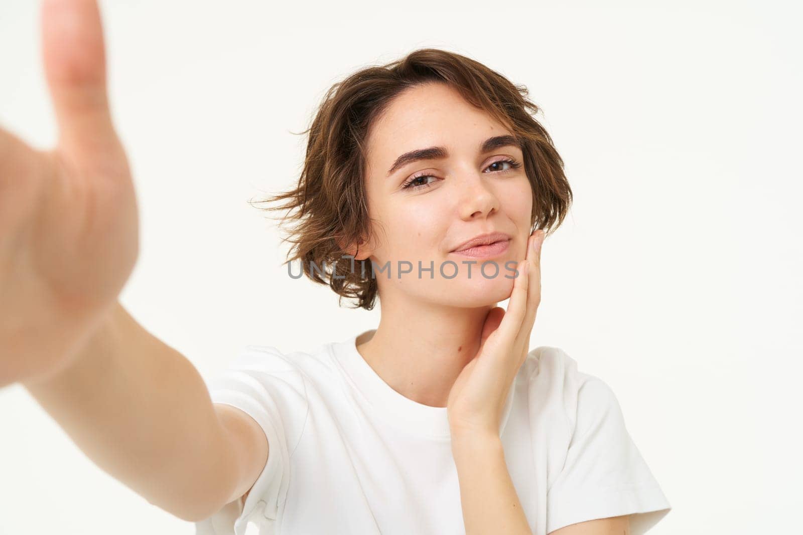 Lifestyle and people. Young woman smiling and taking selfie, posing for photo, holding camera with one hand, standing over white background.