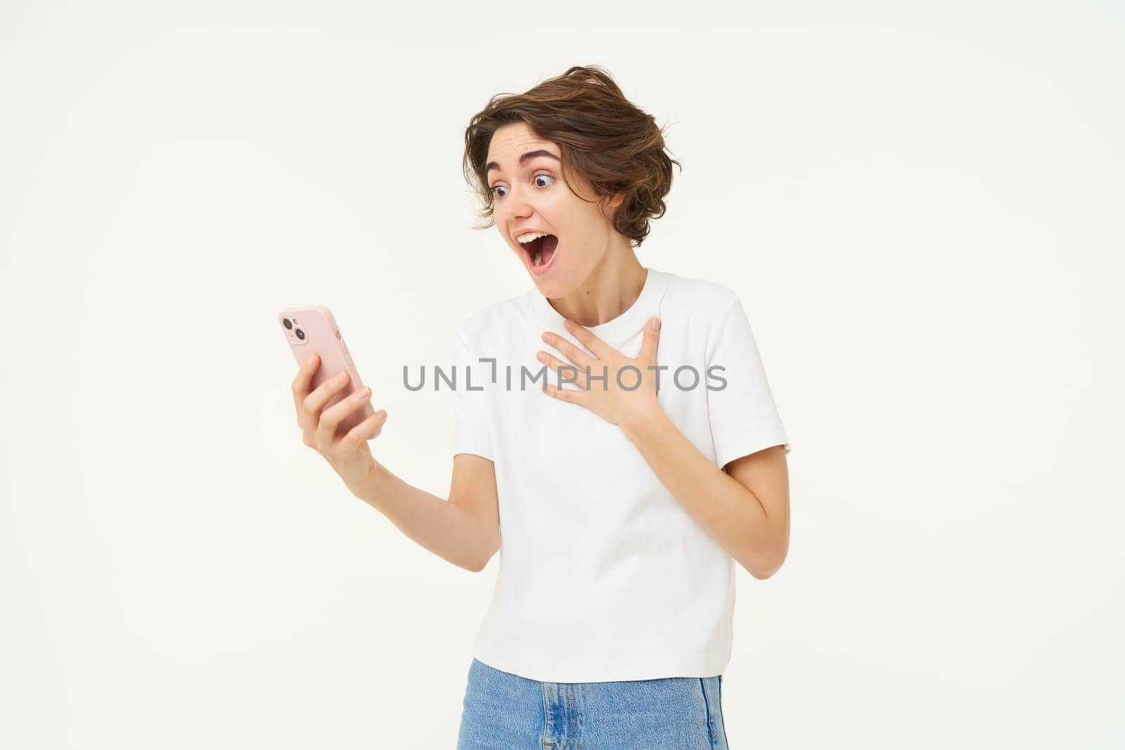 Woman looks at smartphone with happy surprise, reacts to amazing news on mobile phone, reading text with excitement, stands over white background.