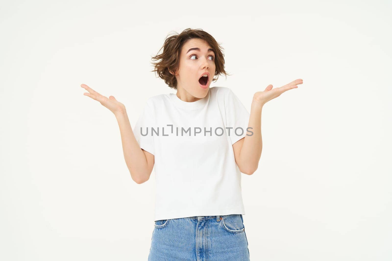 Portrait of brunette woman with surprised face, shrugging shoulders and looking aside with amazed, shocked expression, standing over white background.