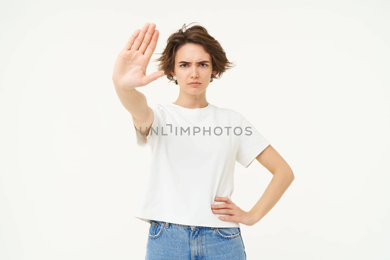Image of serious young woman extends one hand, shows stop, prohibit gesture, forbid something, standing over white background.