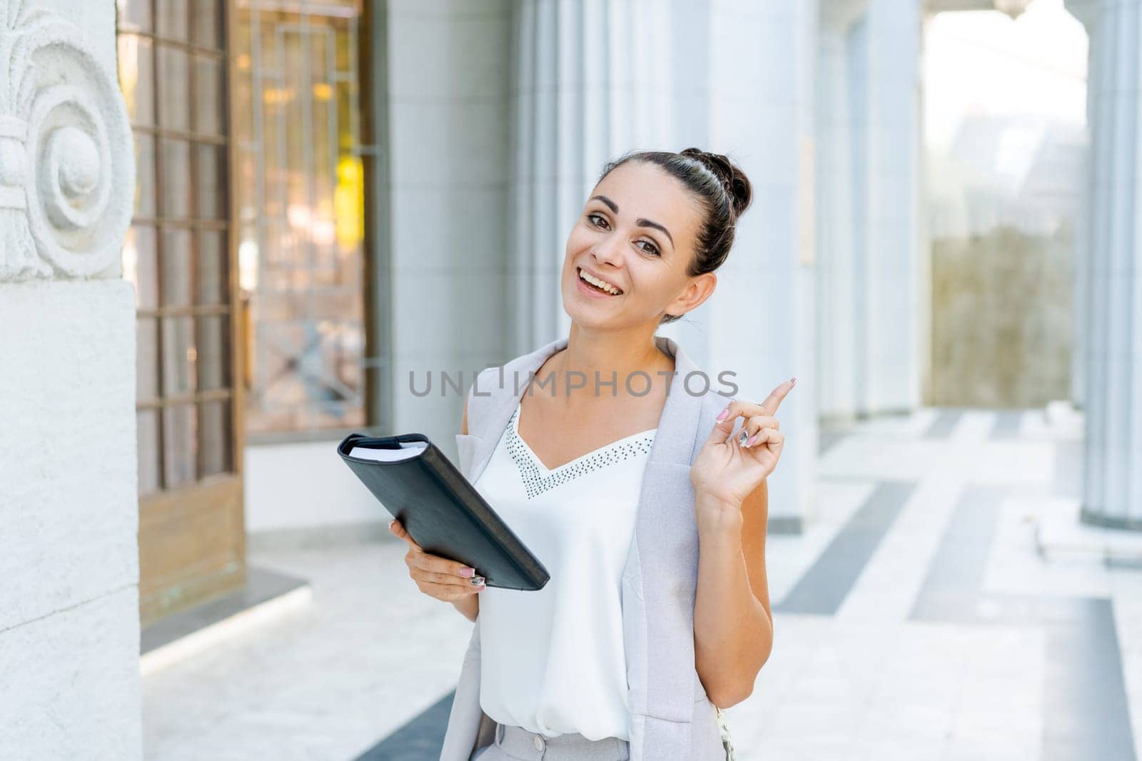 Confident young woman laughing and carrying notepad in hand while standing on the street, wearing stylish clothes