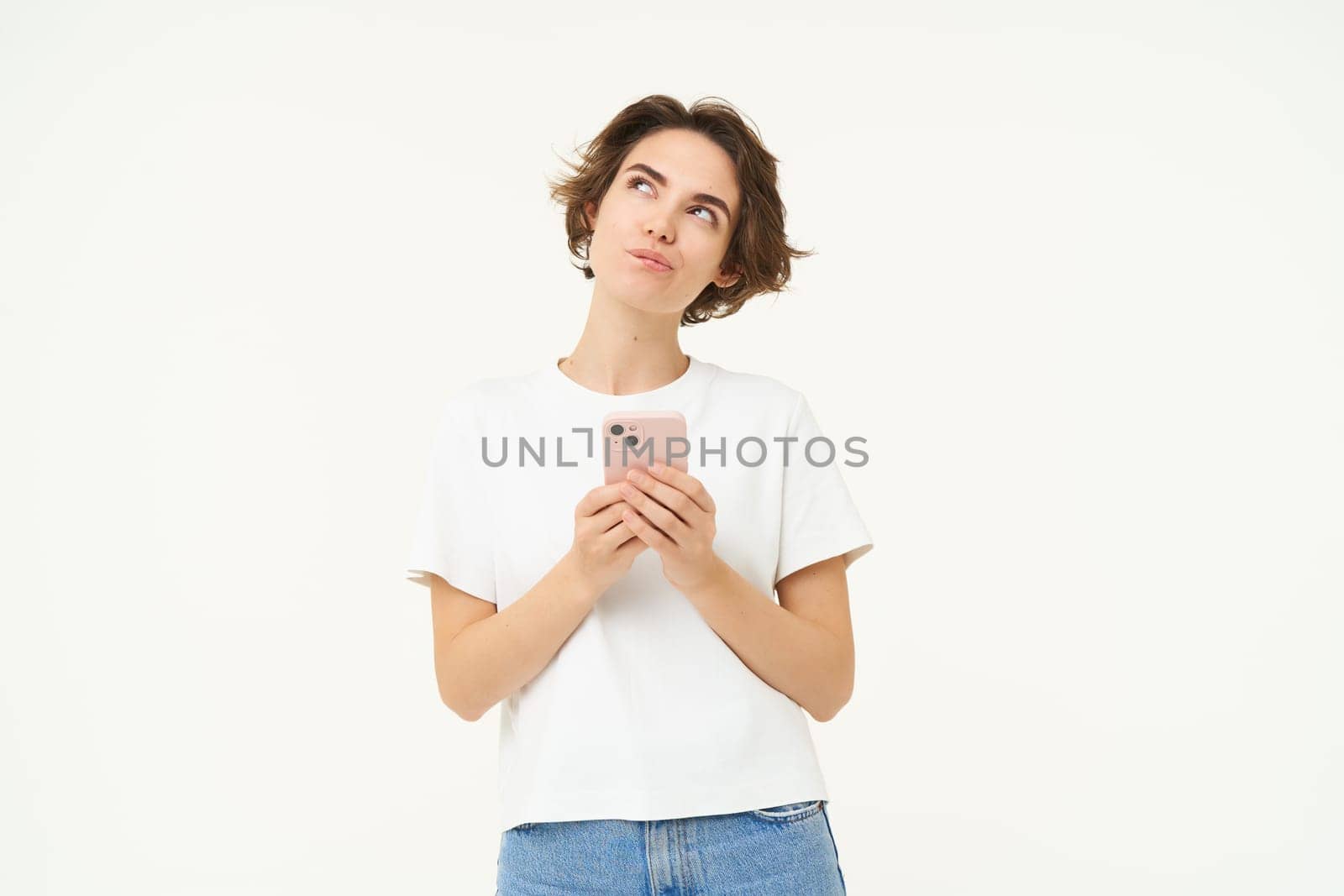 Portrait of thinking girl standing with smartphone, looking up thoughtful, making choice while buying smth online, placing an order, standing over white background.