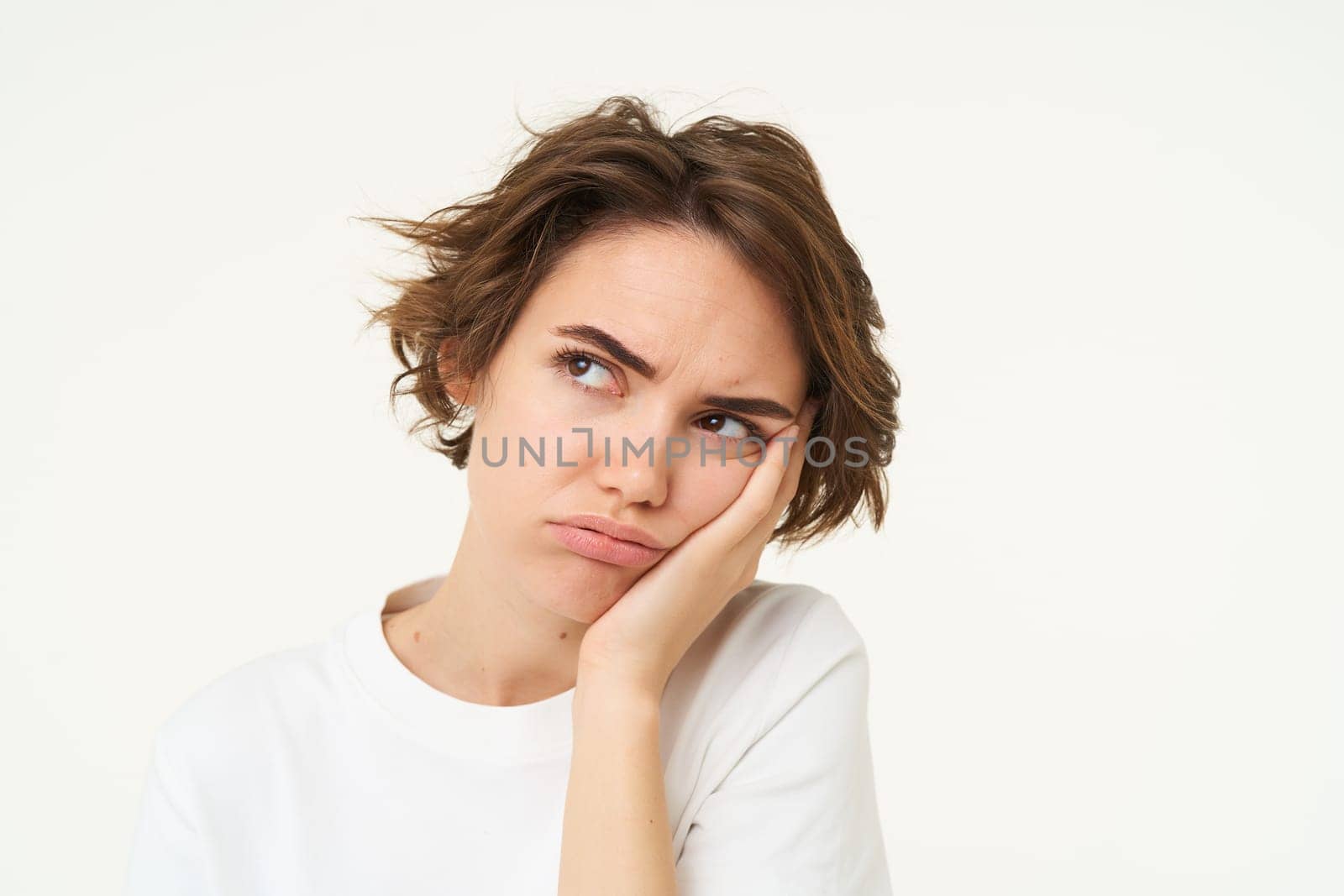 Portrait of young bored woman, leans head on hand and frowns, looks upset and grumpy, thinking, standing over white background.