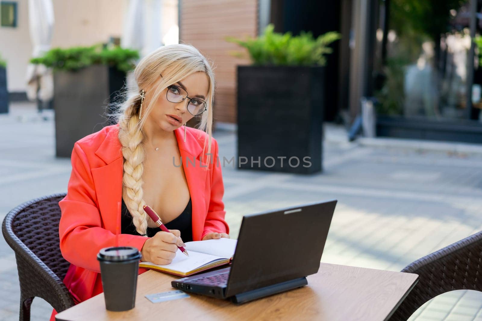 Beautiful woman works on laptop in coffee shop and using mobile phone in a red dress and glasses, business blonde online business