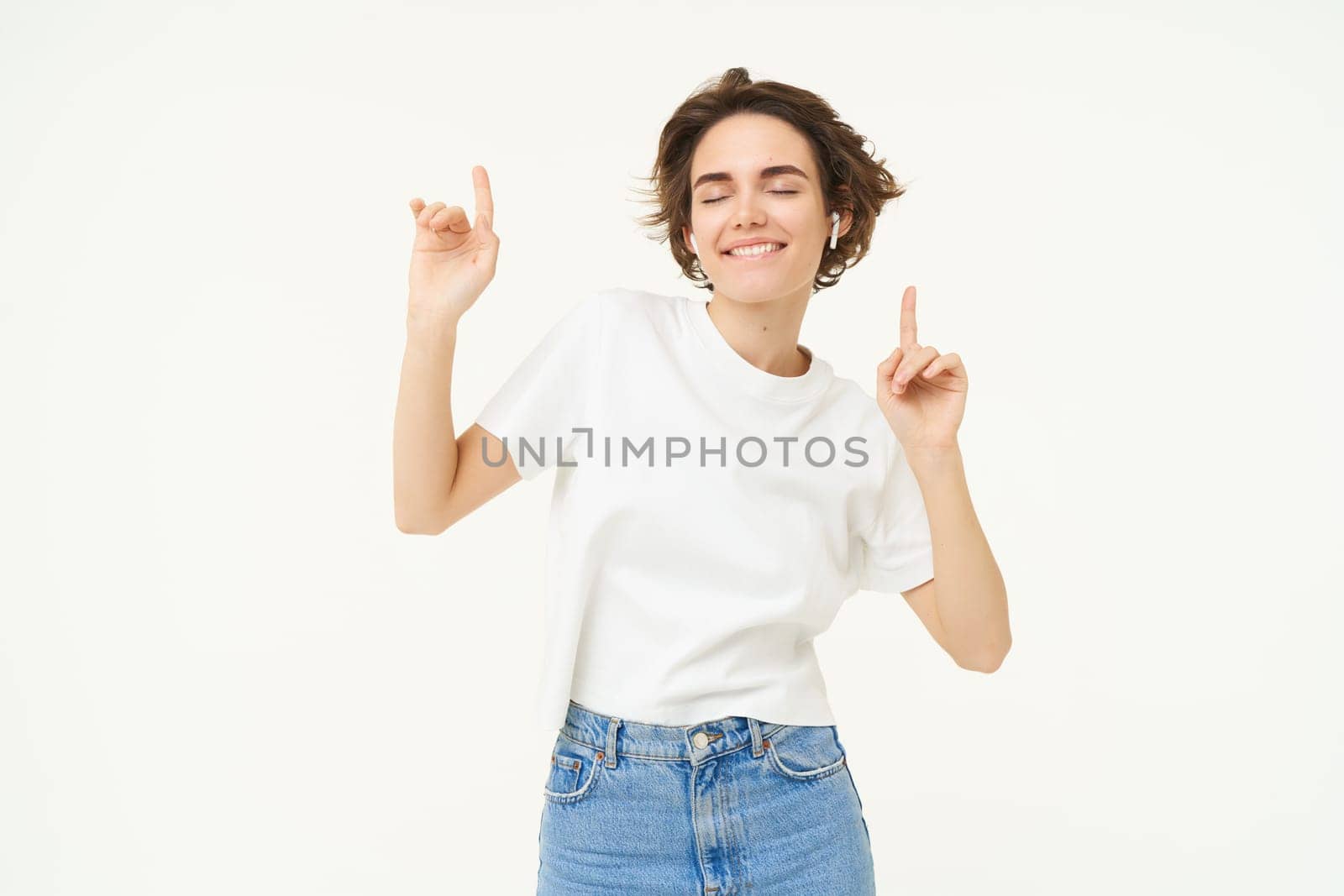 Portrait of happy smiling woman in wireless earphones, dancing and listening to music, enjoying her favourite song, posing over white background.