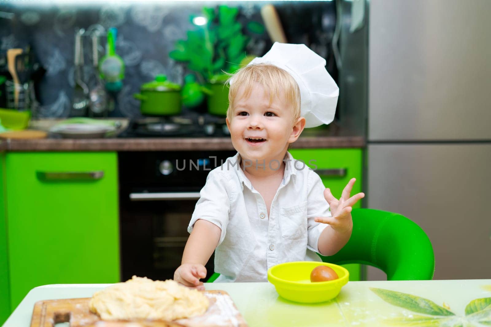 happy little boy preparing dough in kitchen at table. there are dough products on table, dressed as chef by EkaterinaPereslavtseva