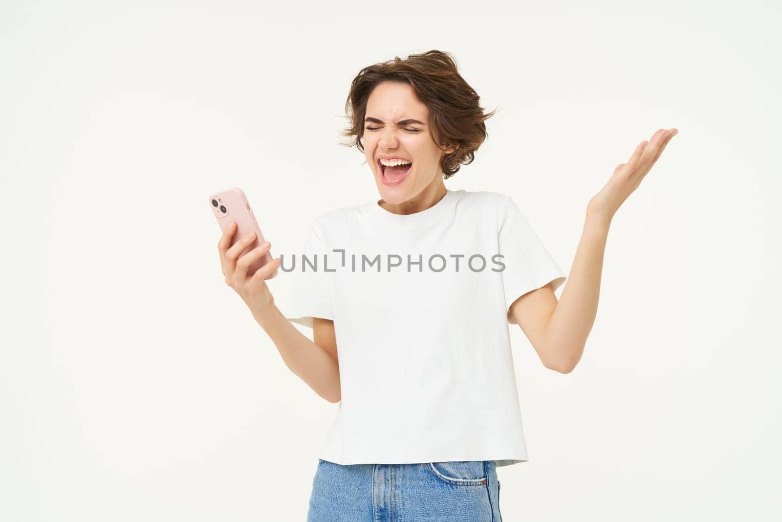 Portrait of woman singing and looking excited, holding mobile phone, triumphing, standing against white background by Benzoix