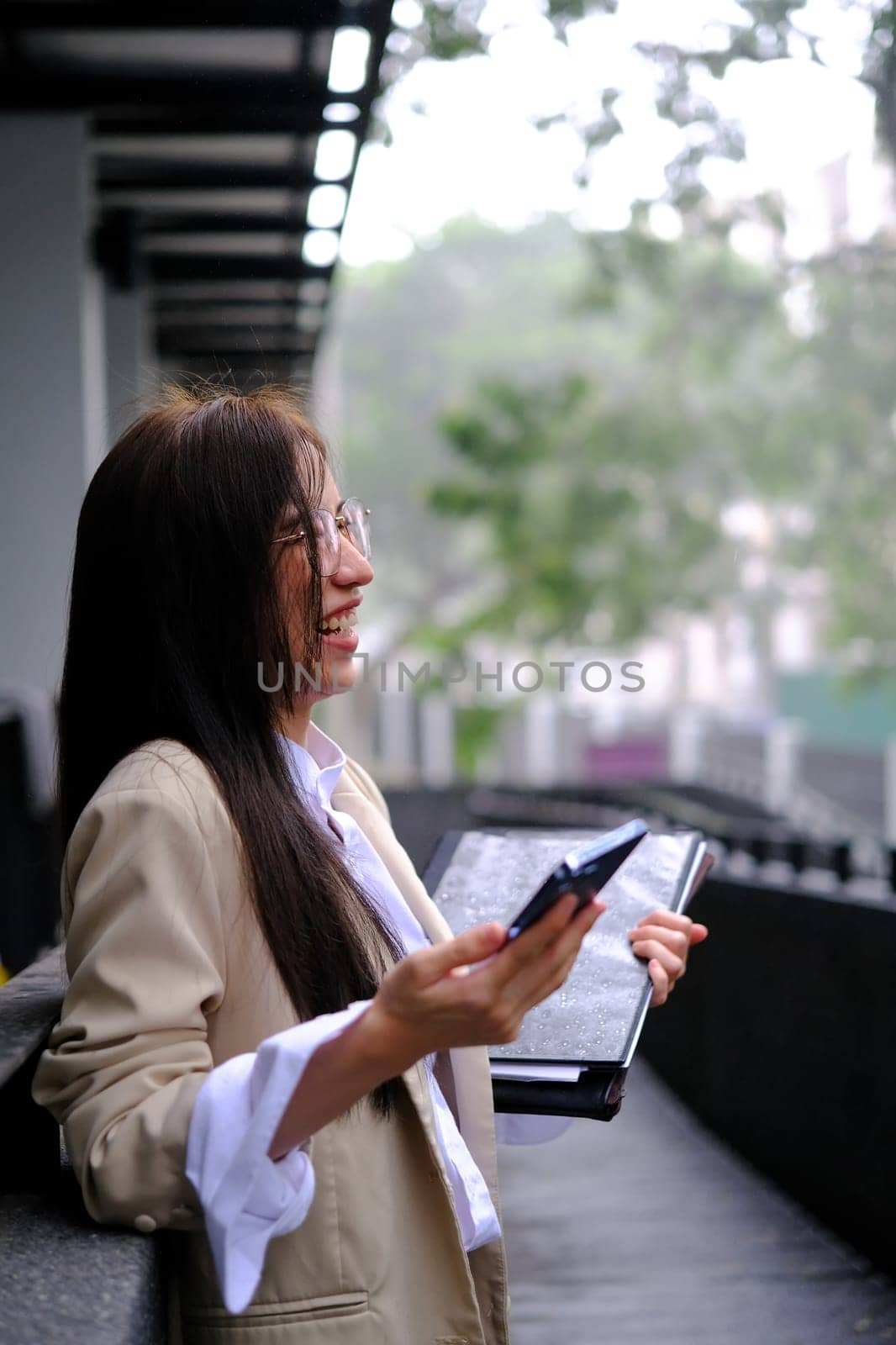 Portrait of cheerful millennial businesswoman holding mobile phone and laughing while standing near office building.