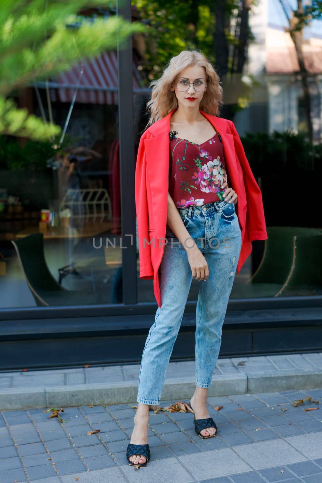 beautiful blonde woman in glasses dressed in red jacket and blue jeans posing on street in the city