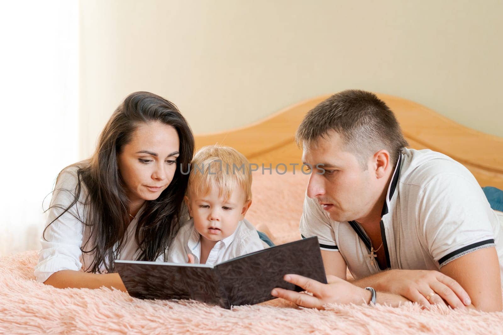 happy family lying on the bed reading a book, mom dad and little son by EkaterinaPereslavtseva