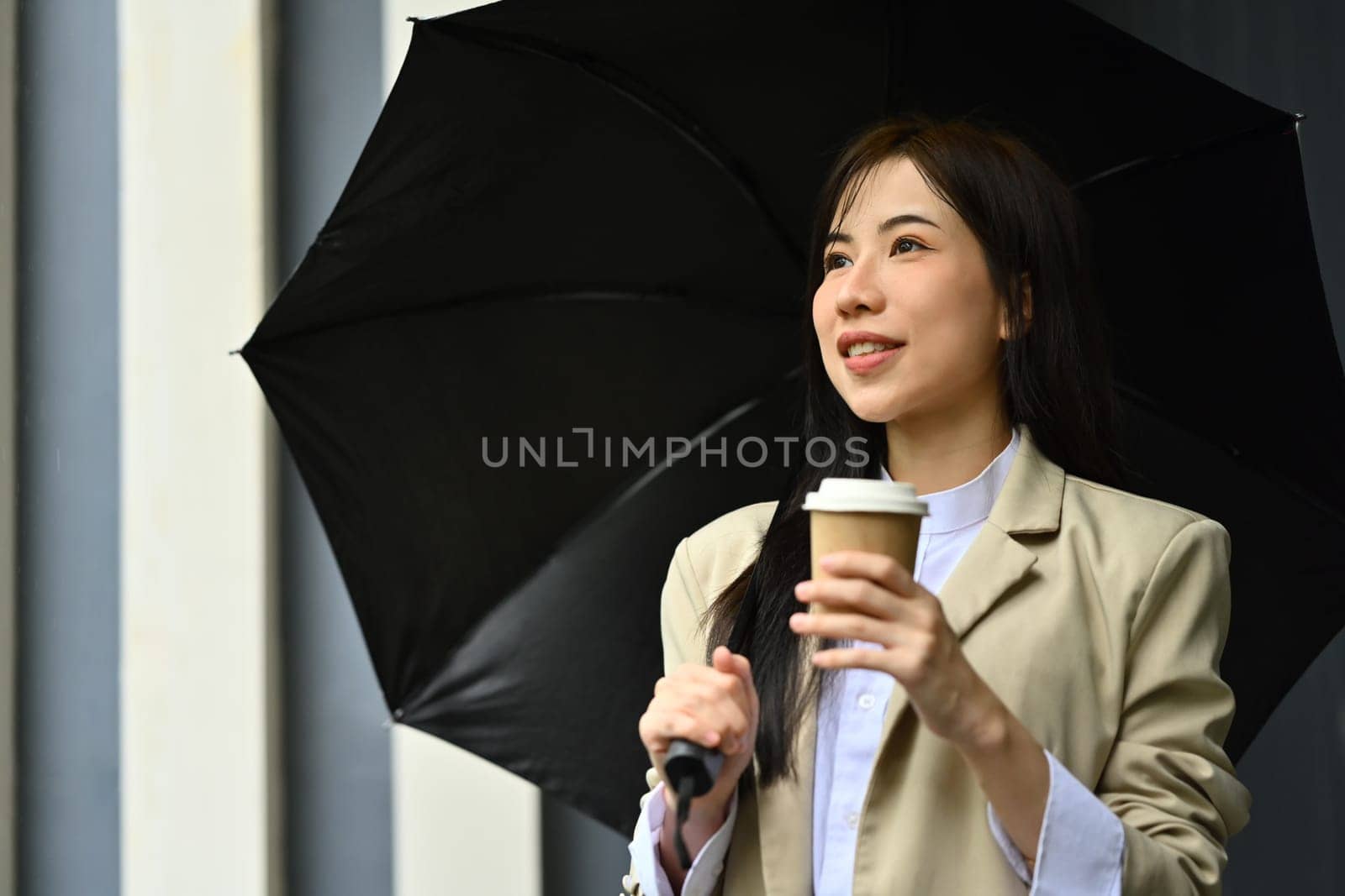 Elegant millennial businesswoman with umbrella and disposable paper coffee cup walking down city street.
